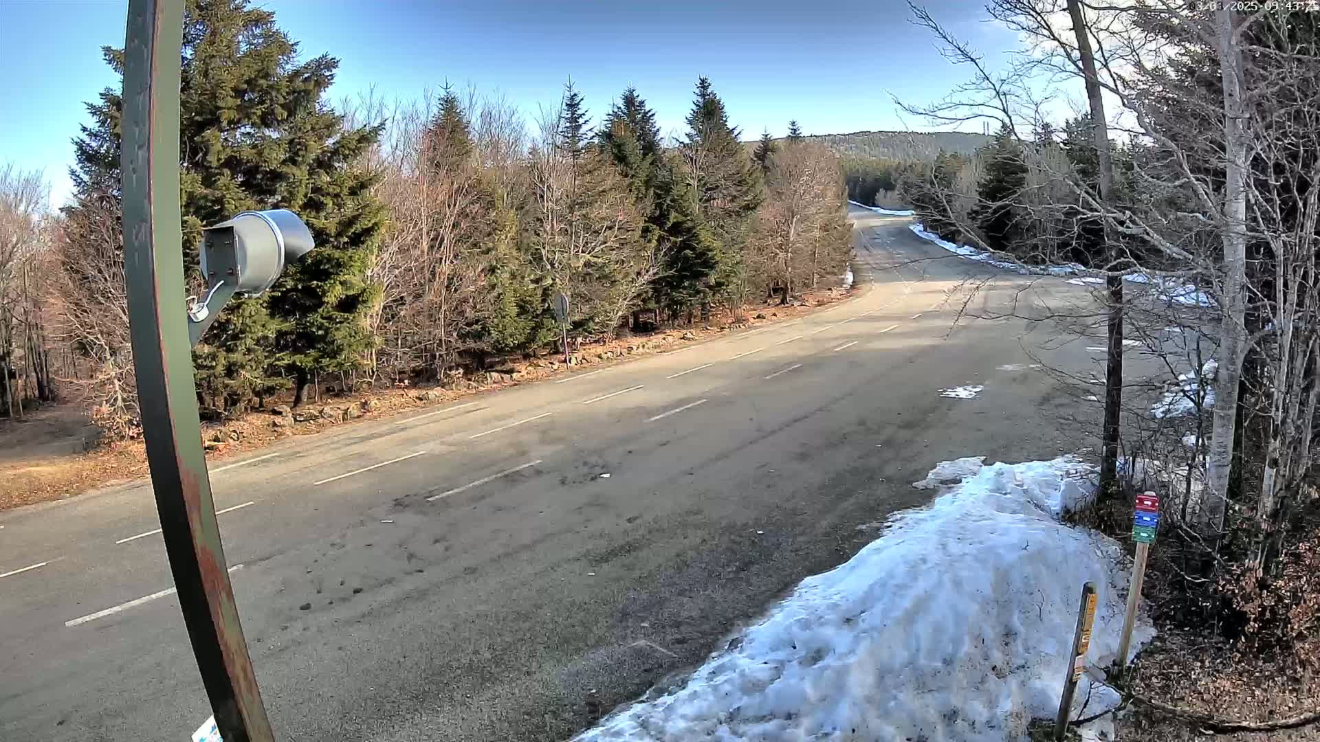 A mostly empty asphalt road curves through a wooded area on a sunny day with patches of snow on the ground.