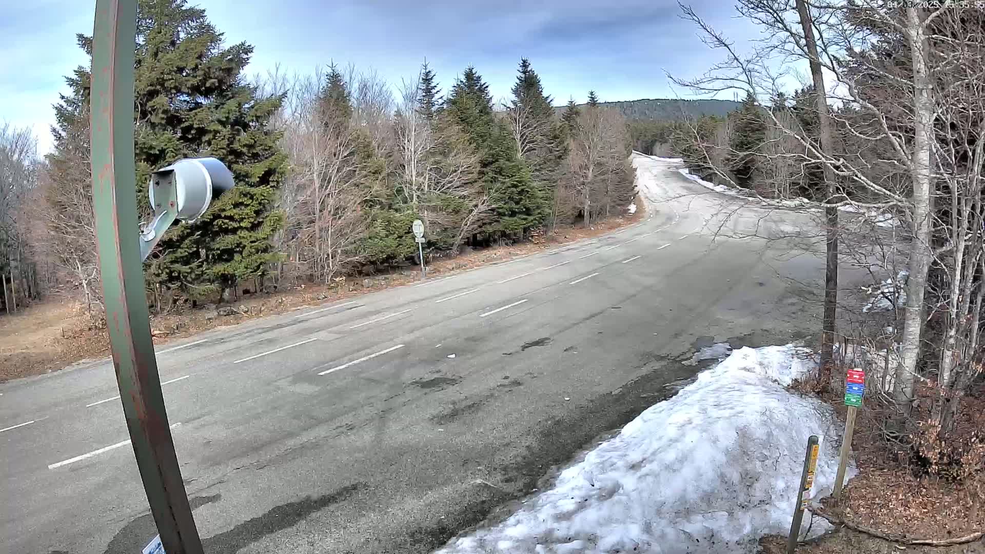 A mostly clear, cool day reveals a paved road intersection in a wintry forest with patches of snow and leafless trees.