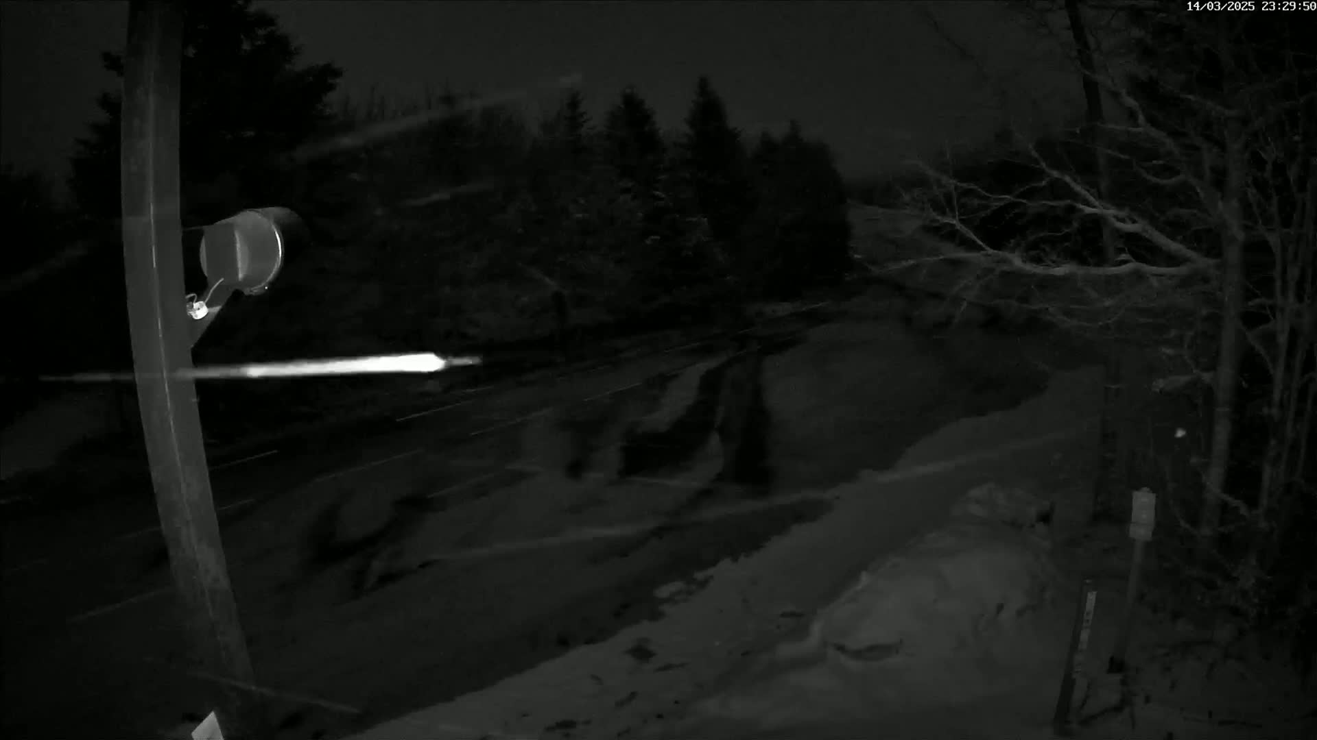 A nighttime, snowy scene shows a road with a vehicle's headlights visible in the distance, viewed from beside a post with a security camera attached.