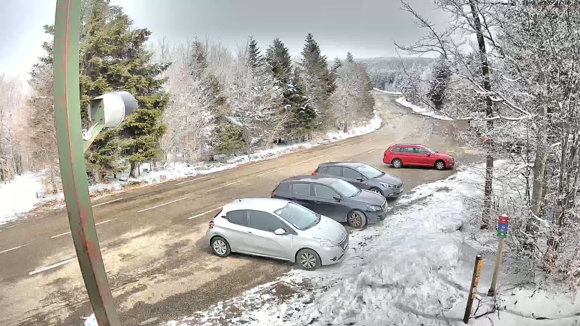 Three cars are parked in a snowy roadside lot next to a winding road bordered by snow-covered trees under an overcast sky.