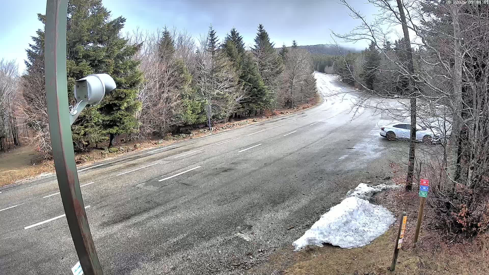 A mostly clear, cool day shows a mountain road curving through a mostly bare forest, with a white car parked on the side, and a small pile of snow near a post.