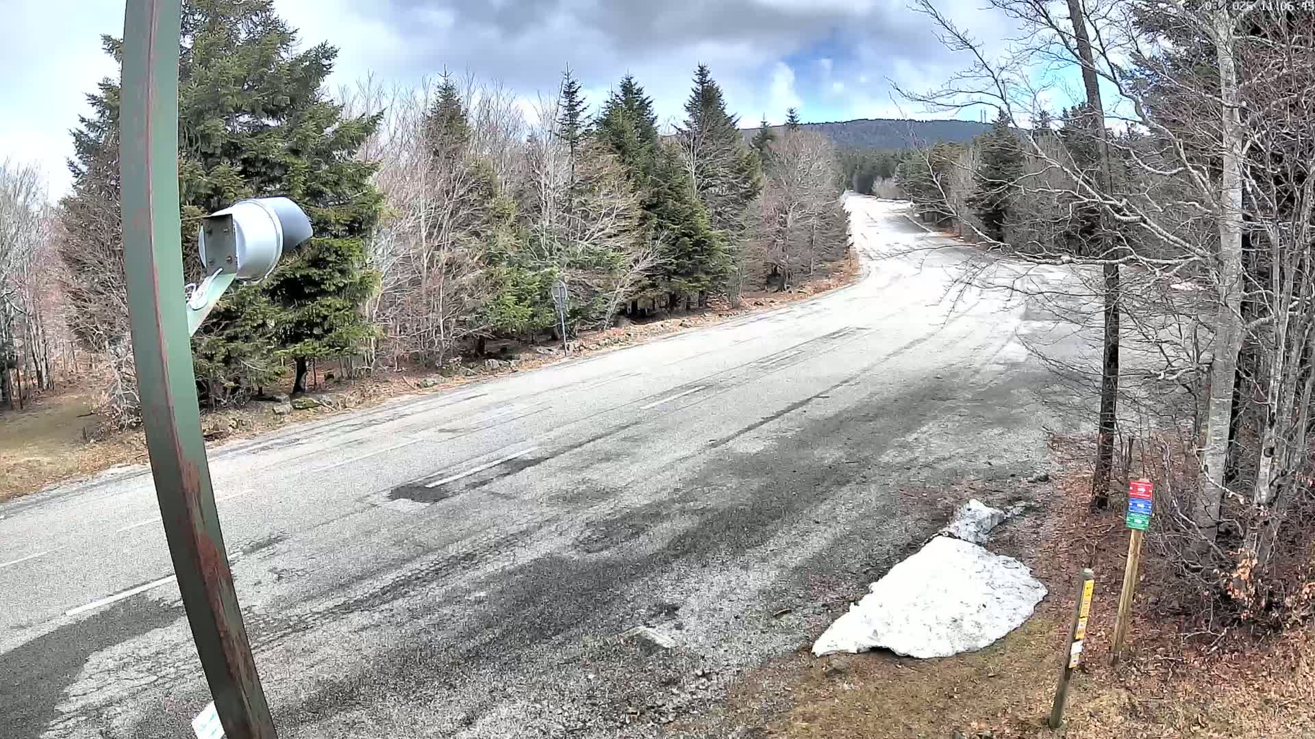 A mostly clear, cool day with patchy snow on the ground shows a winding, empty asphalt road curving through a forest of leafless trees.