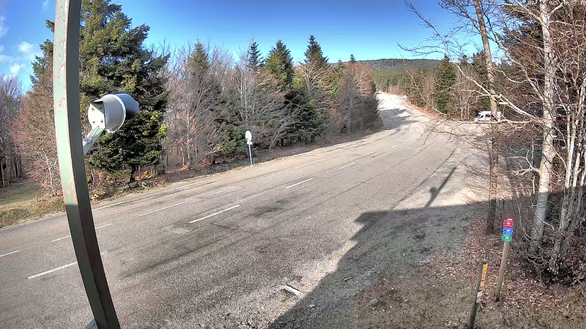 A mostly empty asphalt road curves through a sunny, wooded area with a few bare trees and a single vehicle parked at the side.