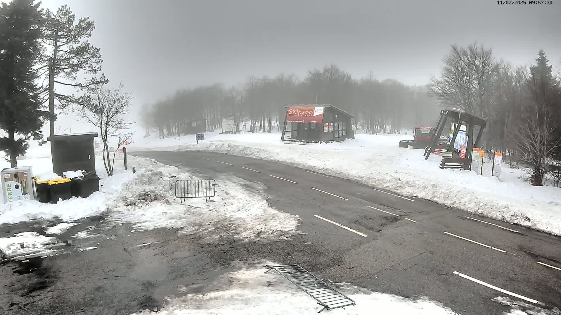 A foggy, snowy scene shows a paved road leading to a small building, with snow-covered ground and trees surrounding it.