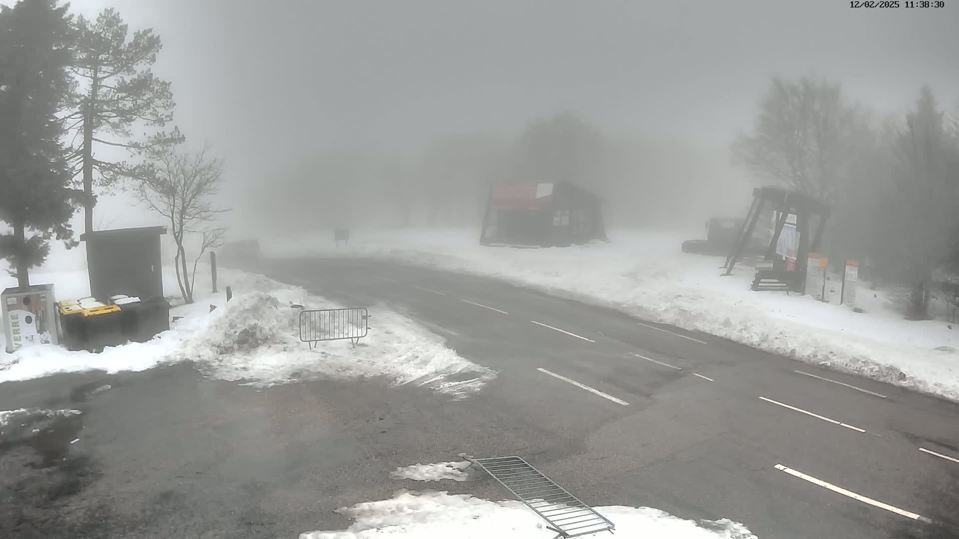 A snow-covered road is shrouded in thick fog, with a few buildings and trees barely visible in the distance.