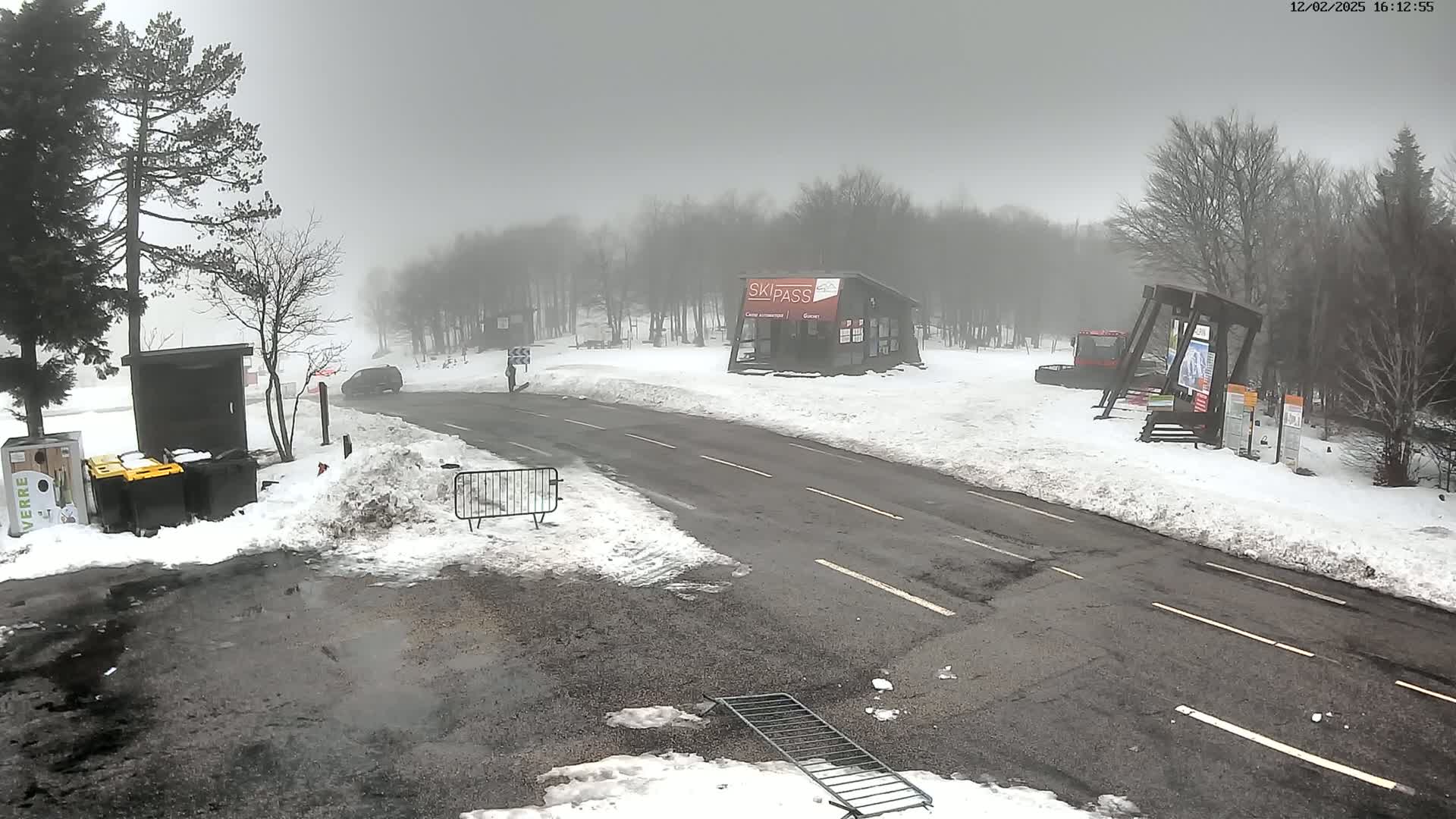 A foggy, snowy scene shows a road passing through a ski resort area with a small building, snow banks, and some equipment.