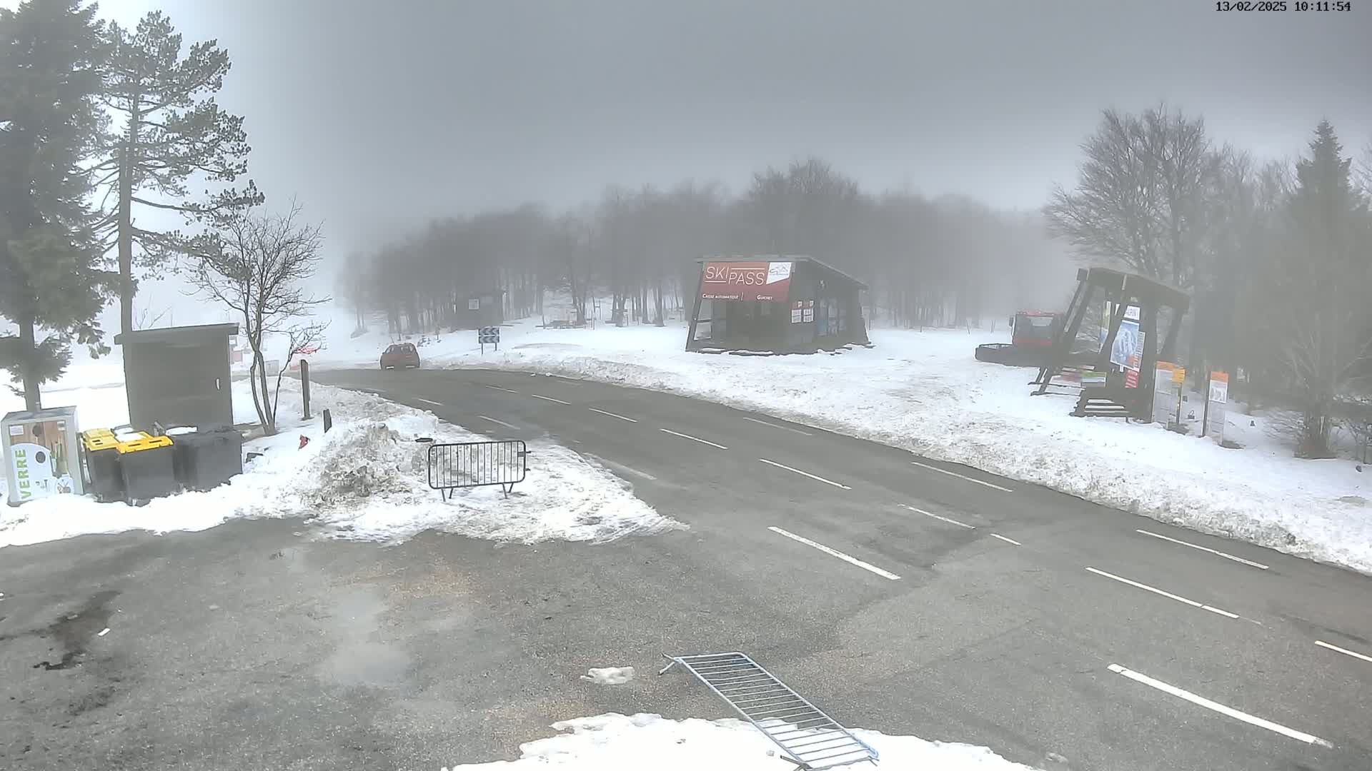 A foggy, snowy scene shows a road leading to a ski pass building, with a few cars and snow-covered ground.