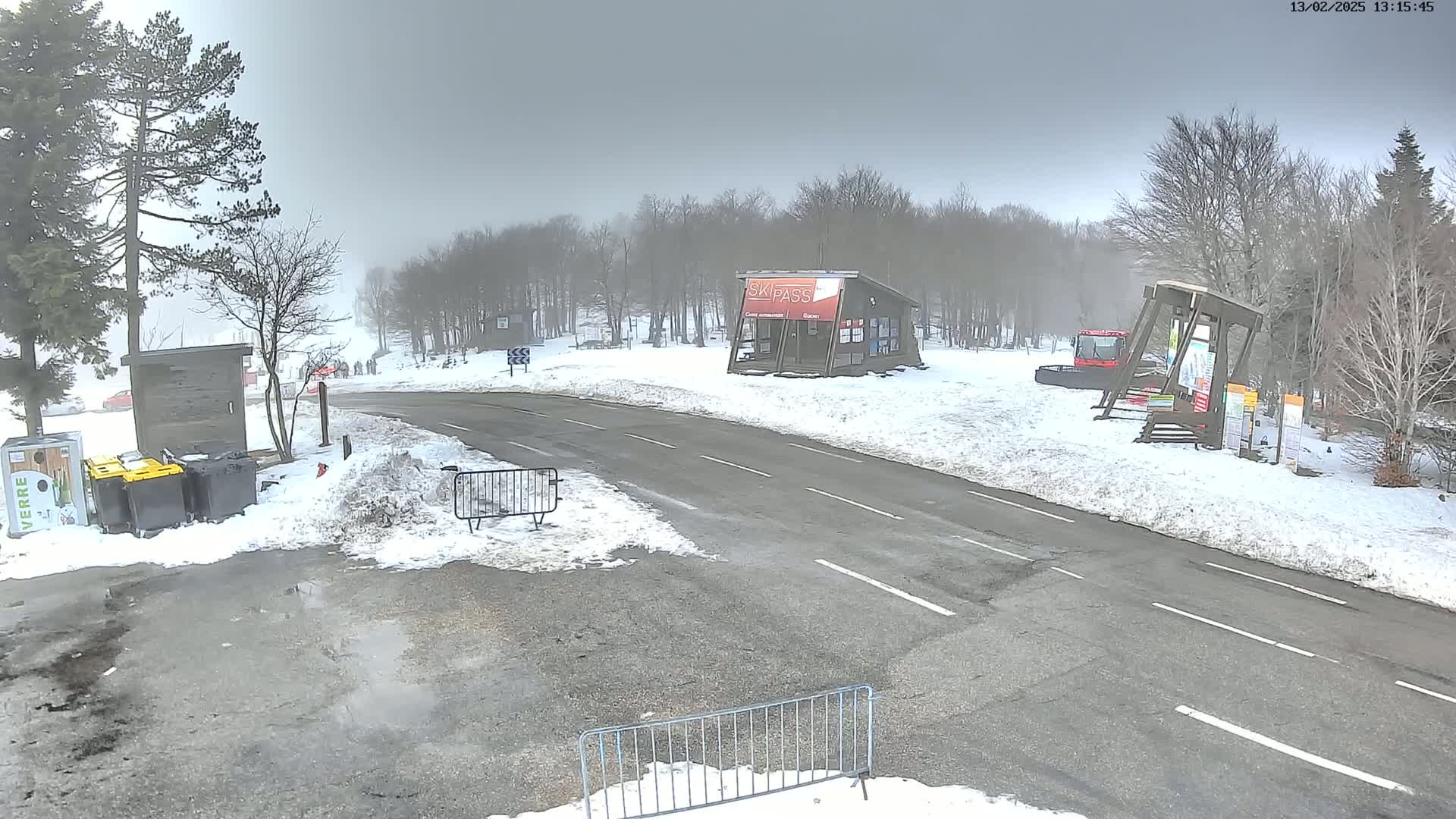 A snowy road curves through a foggy wooded area, where a small building and several trash receptacles are visible near a parking lot.