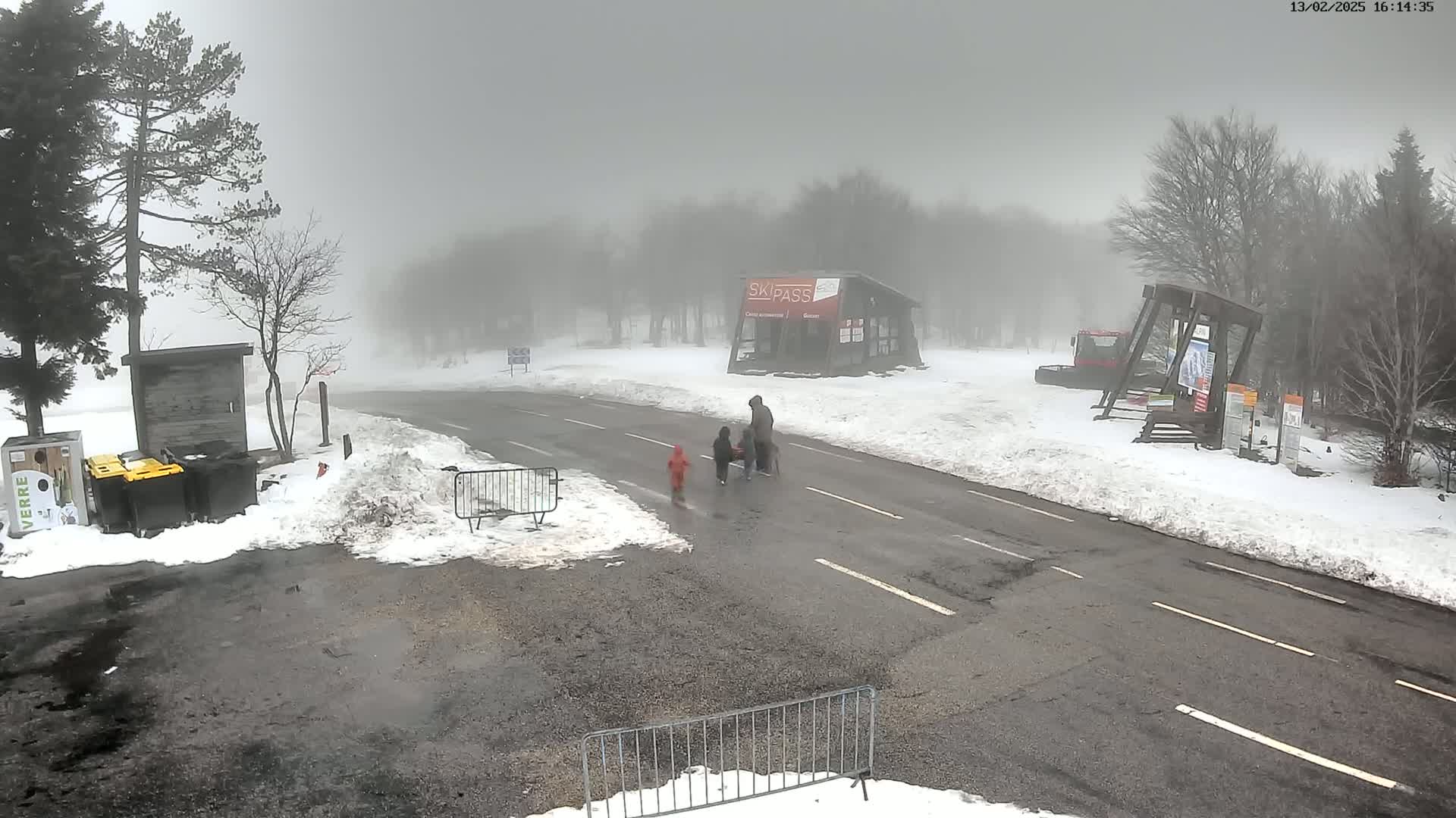 A foggy, snowy scene shows a paved road with a few people walking, some snow-covered ground, and several trees and buildings in the background.