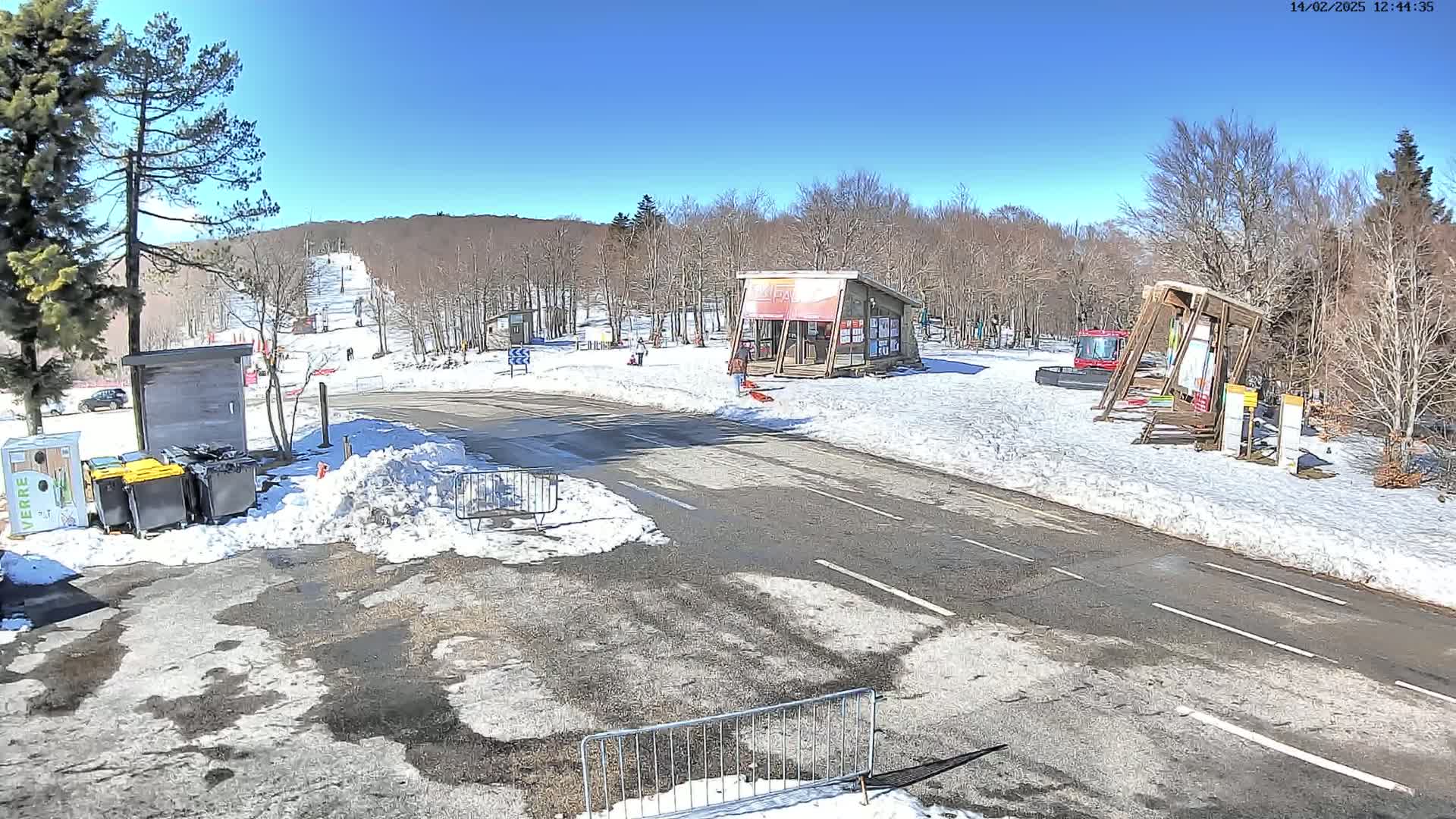 A sunny day at a ski resort shows a paved road with snow on the sides, several small buildings, and a ski lift in the background.