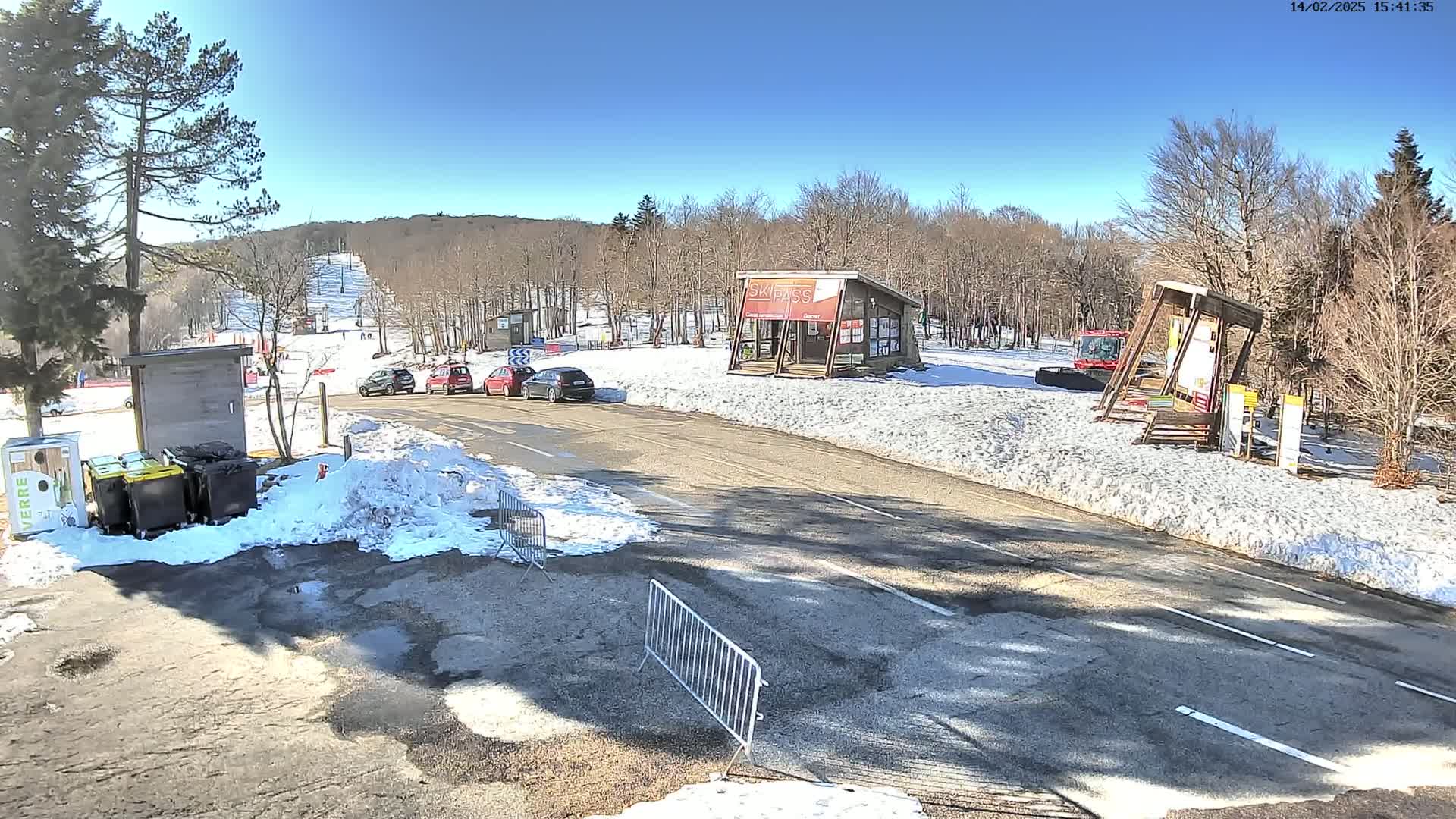 A sunny winter scene shows a ski resort parking lot with several cars parked near a small building and snow-covered ground.