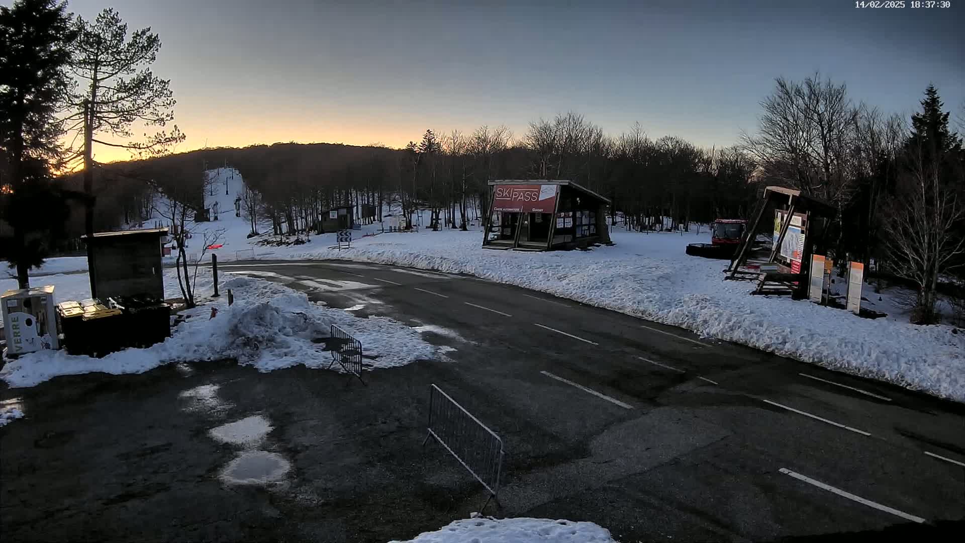 A snow-covered ski resort parking lot at sunset, with a small building and ski lift visible in the background.