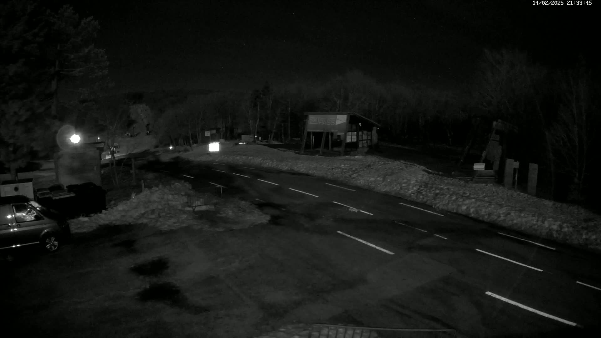 A nighttime, snow-covered road scene shows a van parked near a small building, with a road curving away into a dark, wooded area under a starlit sky.