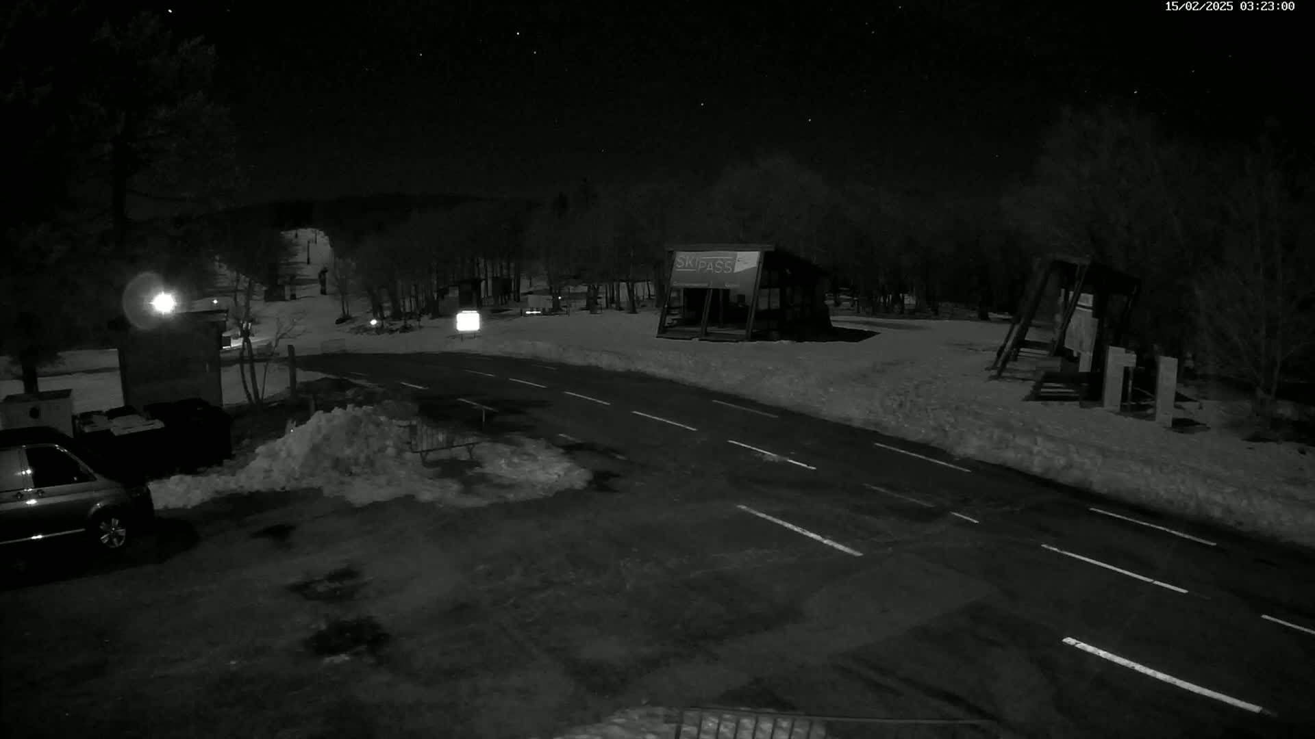 A nighttime, snow-covered parking lot and road scene features a small building, some playground equipment, a van, and a starry sky.