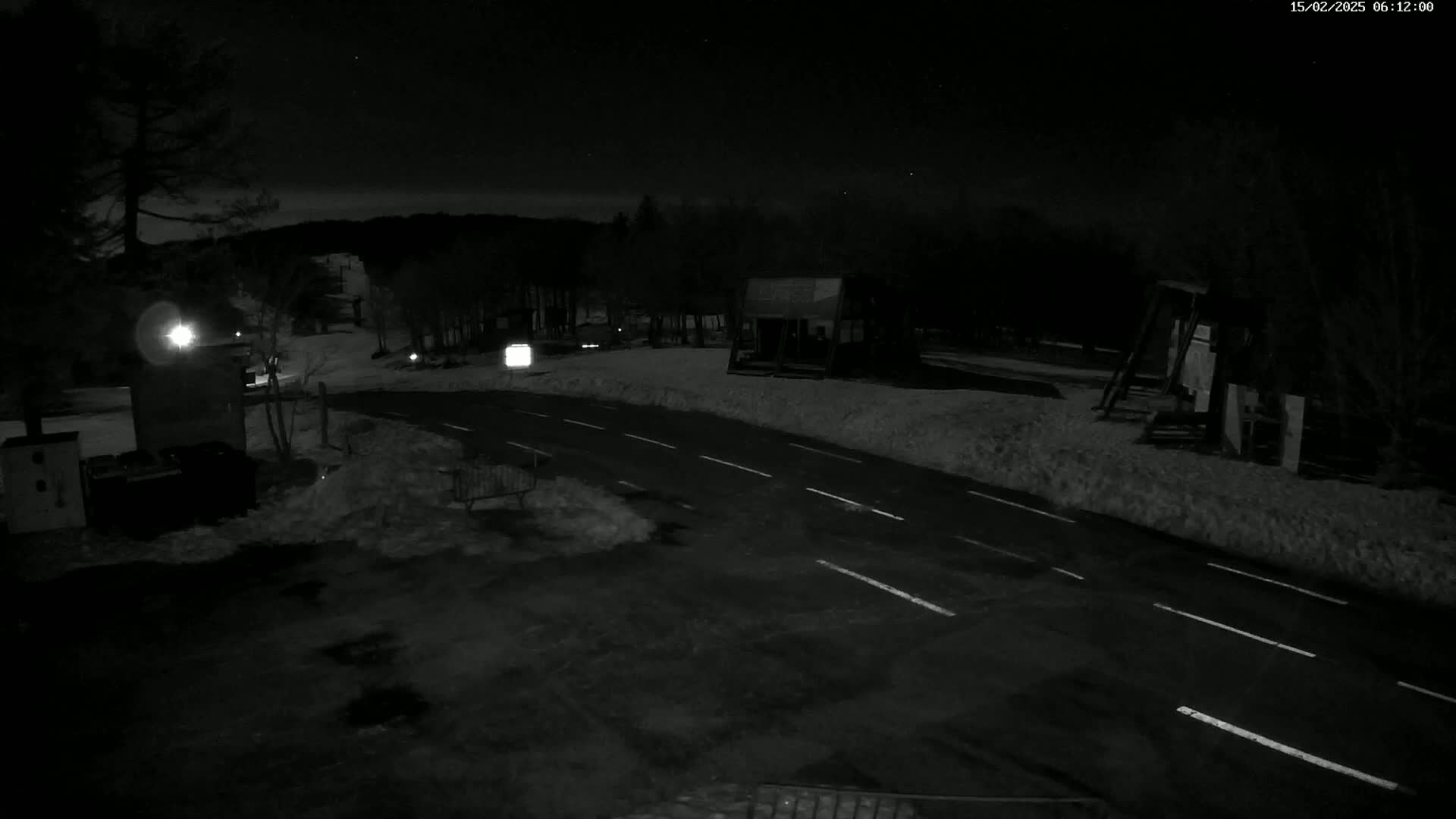 A snow-covered nighttime scene shows a paved road curving through a sparsely lit area with some structures and trees.