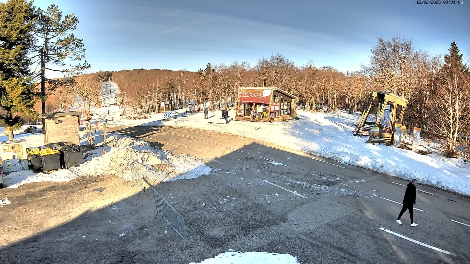 A sunny winter scene at a ski resort shows a parking lot, a ski lift in the background, and a few people near a ticket booth.