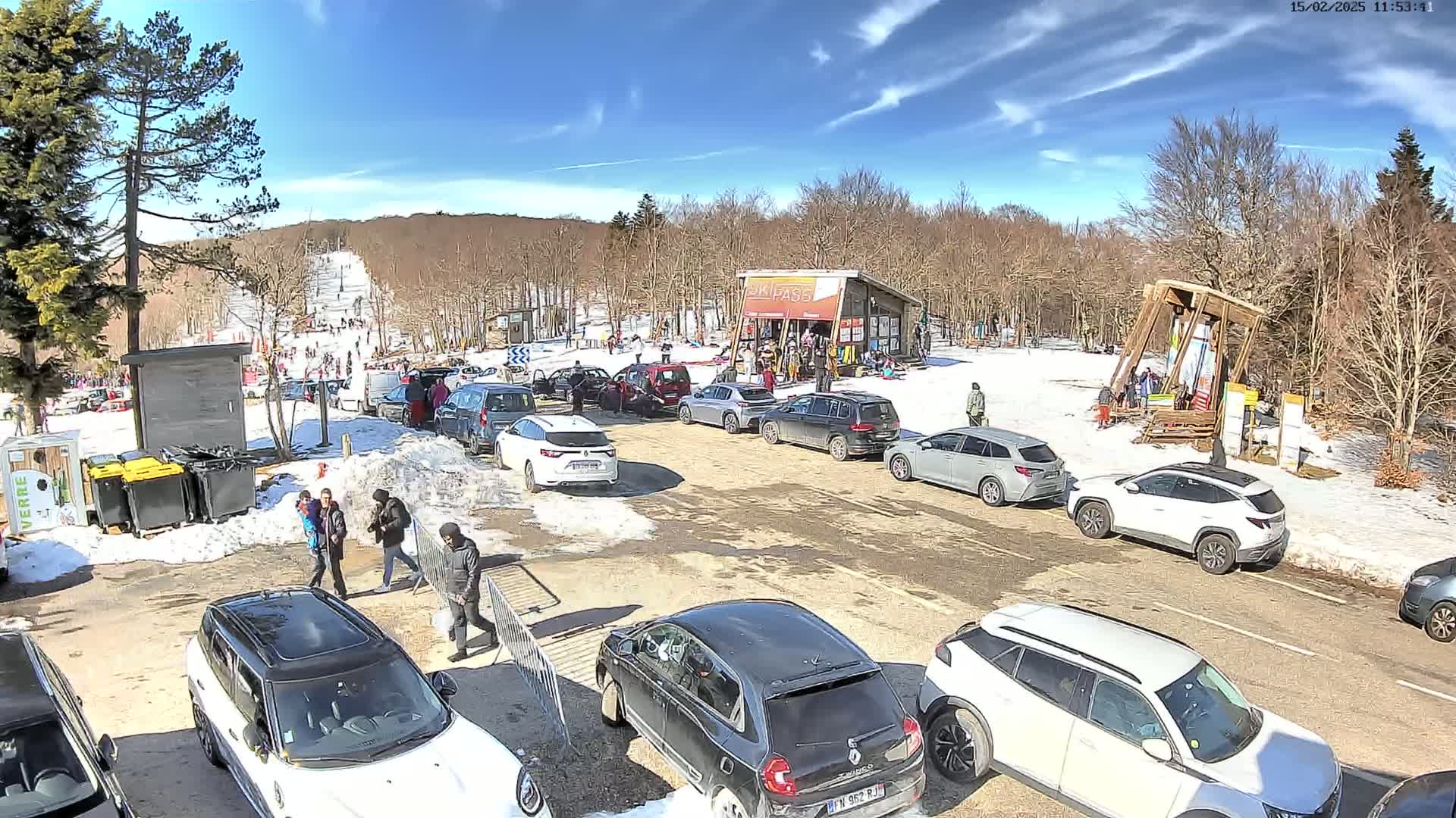 A sunny day at a ski resort parking lot shows numerous cars parked near a small building and ski slope with people visible on the hill and in the parking area.