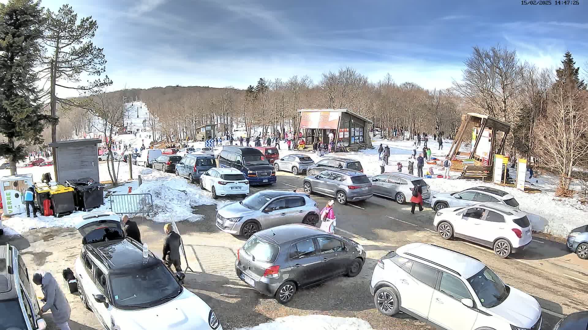 A parking lot full of cars is situated near a ski slope with people on it, under a partly sunny sky.