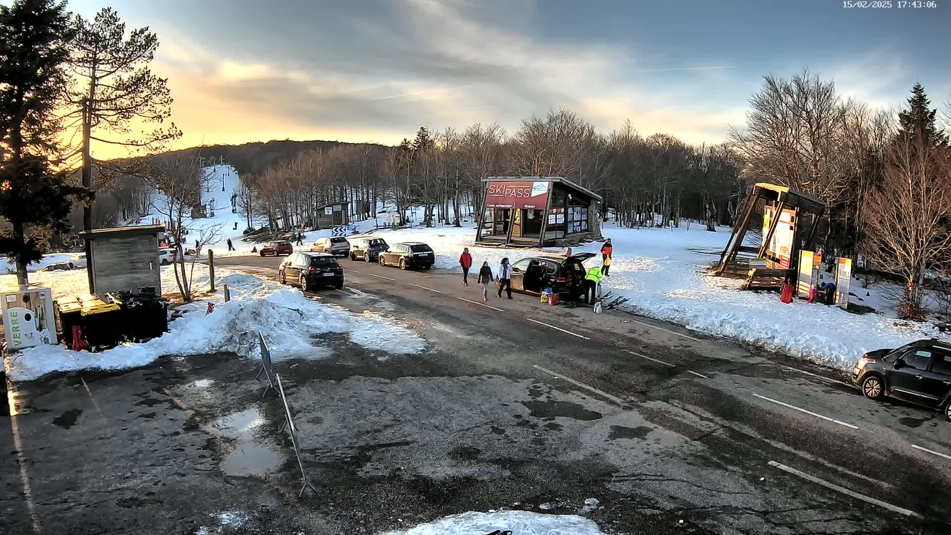 A parking lot and road are partially covered in snow at sunset, with several cars parked nearby, a ski lift visible in the background, and several people walking near a ski pass building.