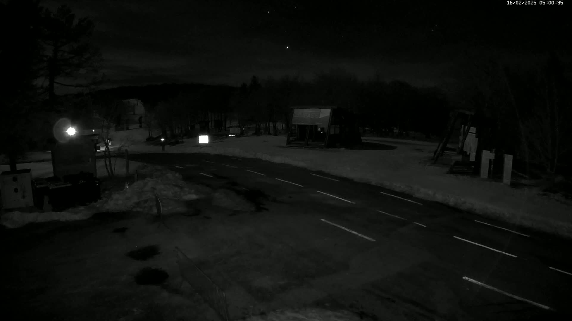 A nighttime, snow-covered road with a few structures and trees under a partly cloudy sky.