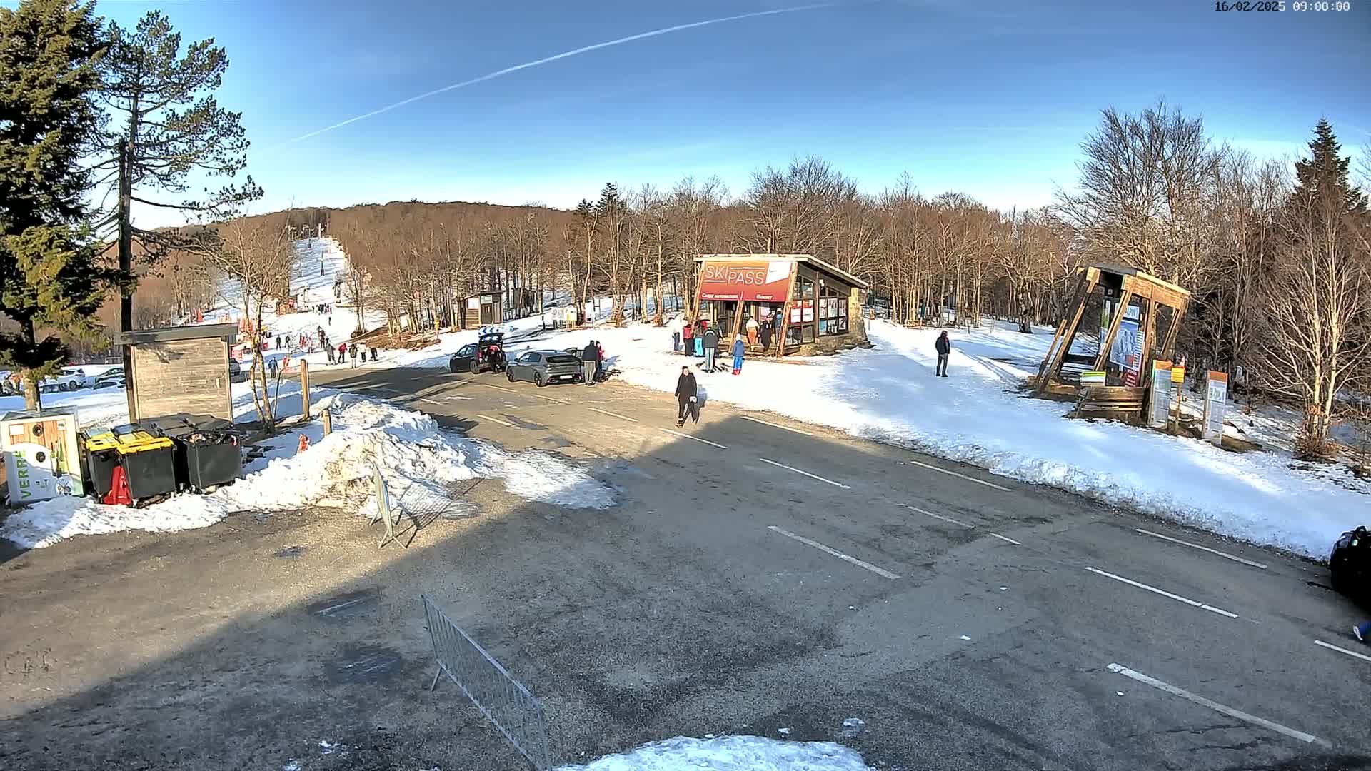 A sunny winter scene at a ski resort shows a parking lot, a ski lift in the distance, several people walking near a ski pass building, and cars parked in the lot.