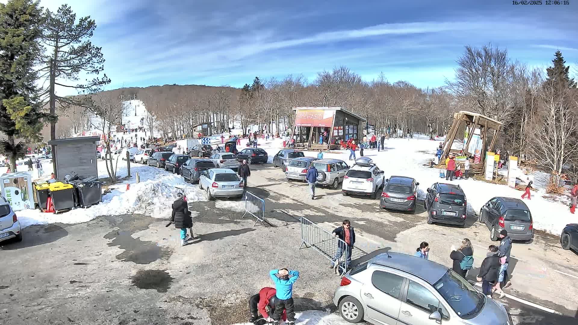 A parking lot with numerous cars is situated near a ski slope on a sunny day with patches of snow on the ground.