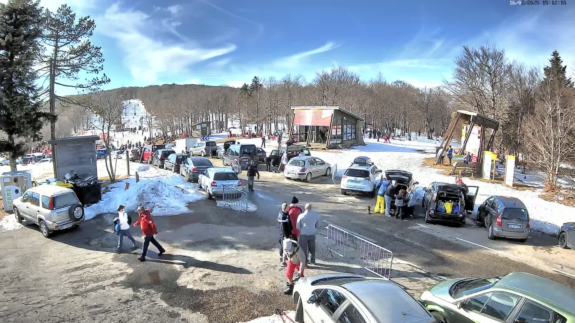 A parking lot with numerous cars is situated at the base of a snow-covered ski slope under a sunny sky.