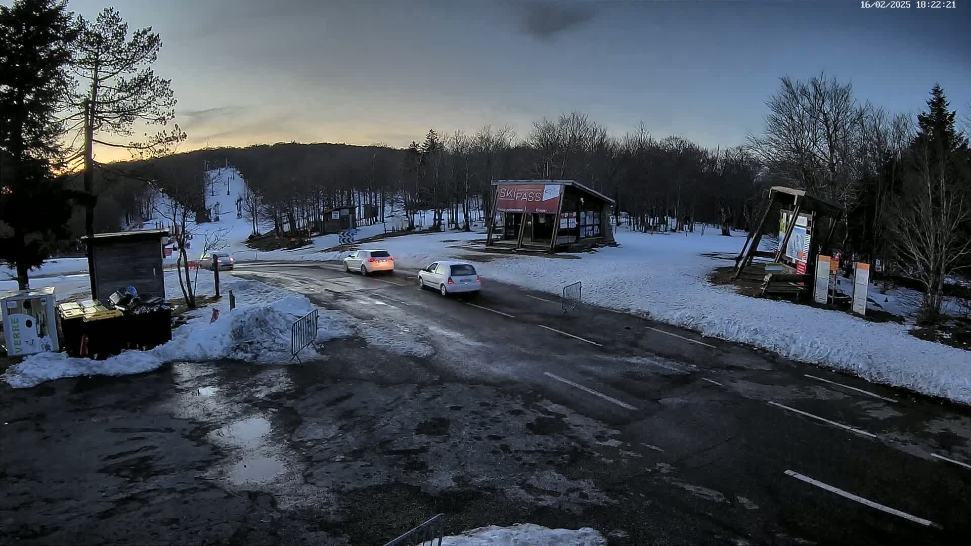 A snow-covered ski resort parking lot at dusk, with a few cars on a road, shows a small building and some trees in the background.