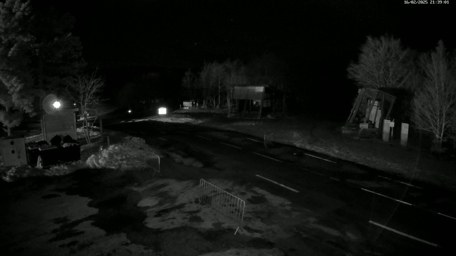 A dark, nighttime scene shows a snow-covered road with small structures and trees on either side, under a clear sky.