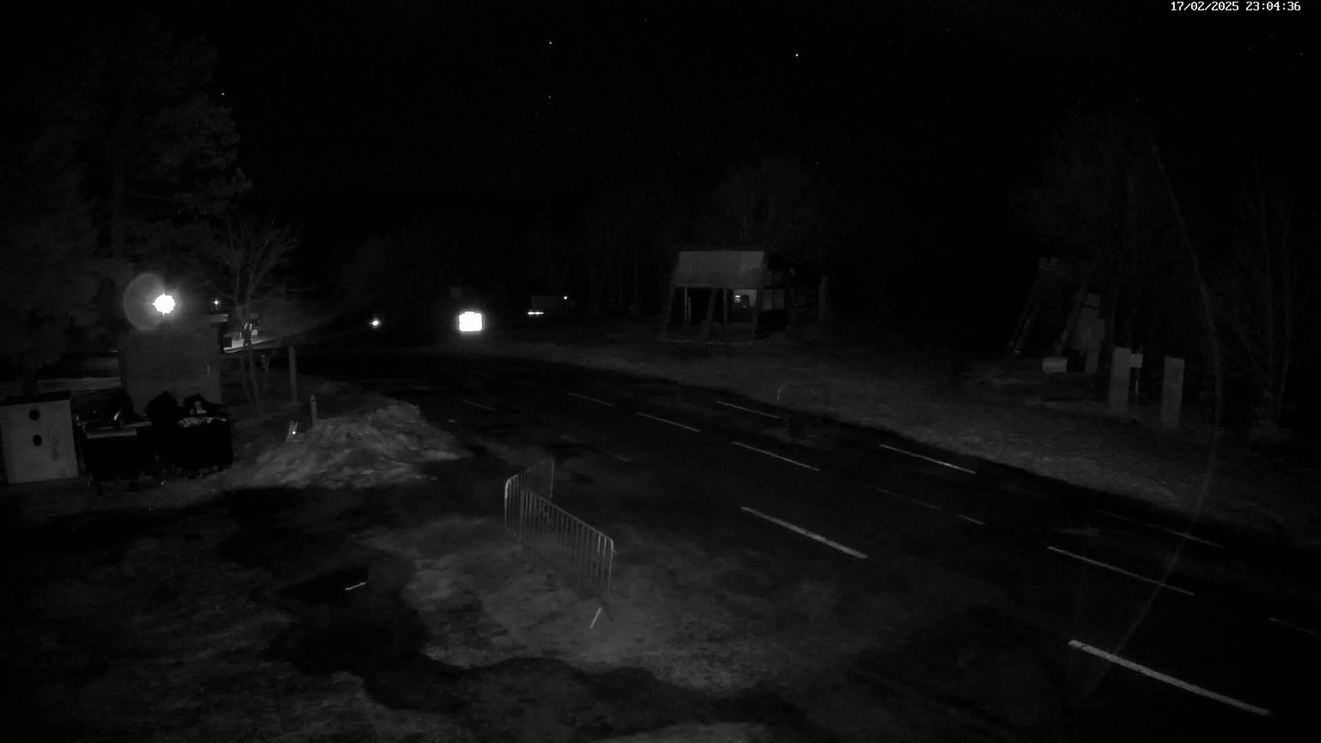 A dark, nighttime scene shows a paved road with faint white lines, bordered by snow and trees, under a clear, starlit sky.