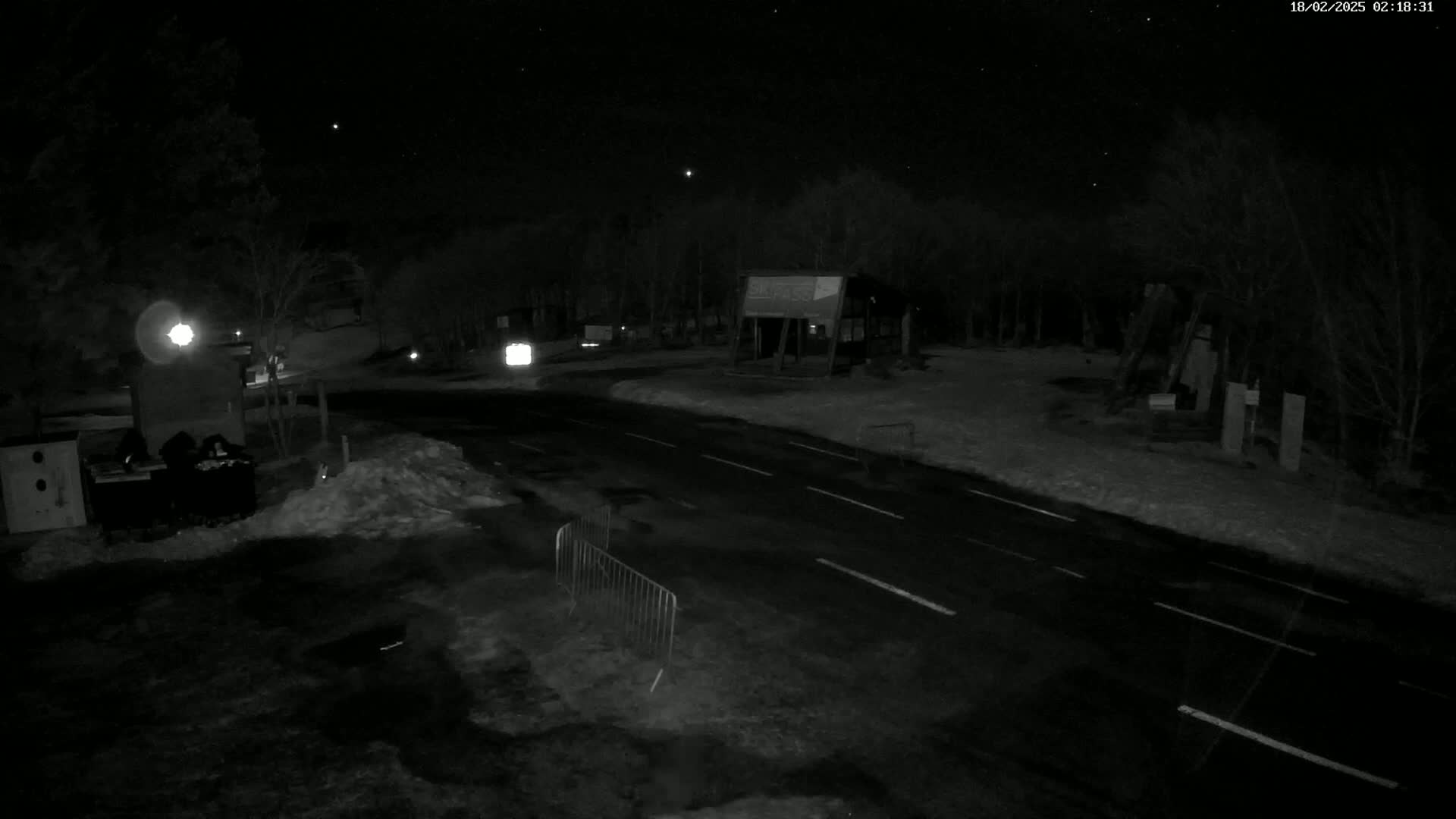 A nighttime, snow-covered roadway curves through a wooded area, illuminated by artificial lights, near a structure that appears to be a small shelter or building.