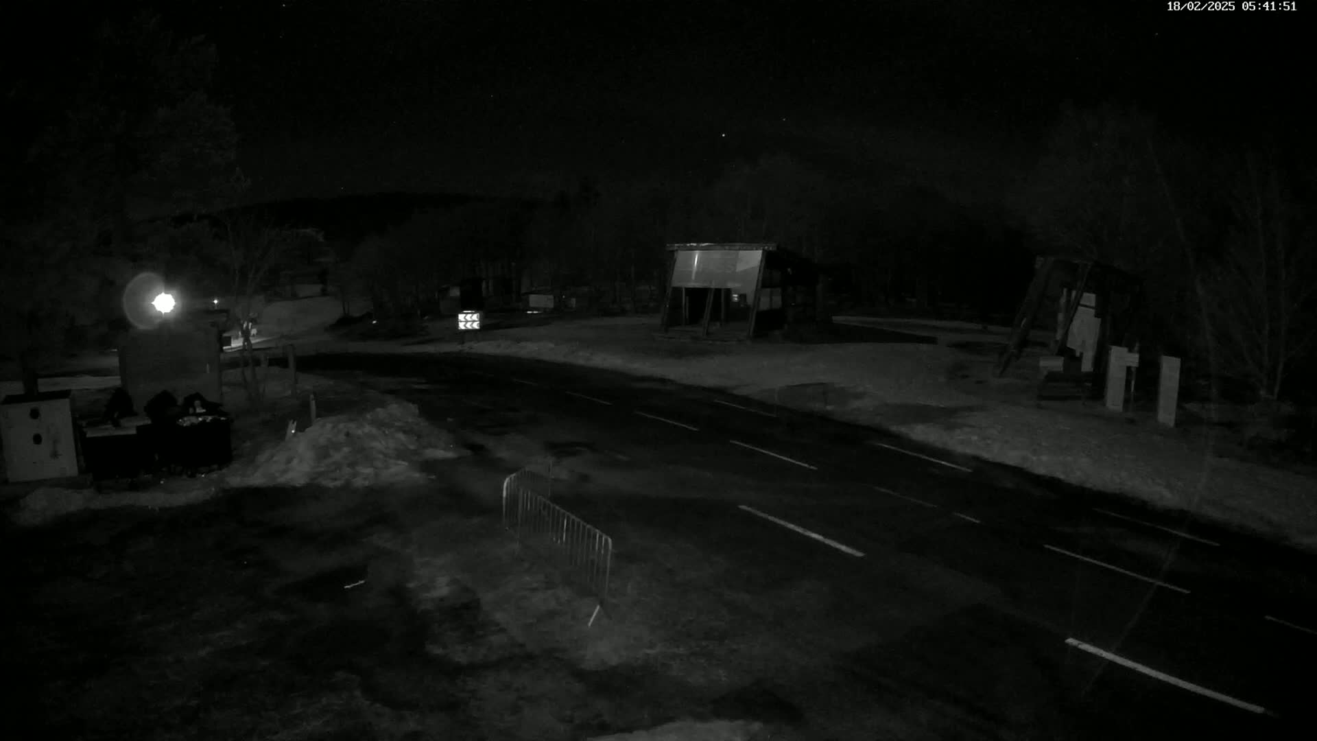 A dark, nighttime scene shows a paved road with lane markings, bordered by snow-covered ground and sparsely lit structures under a clear, starry sky.