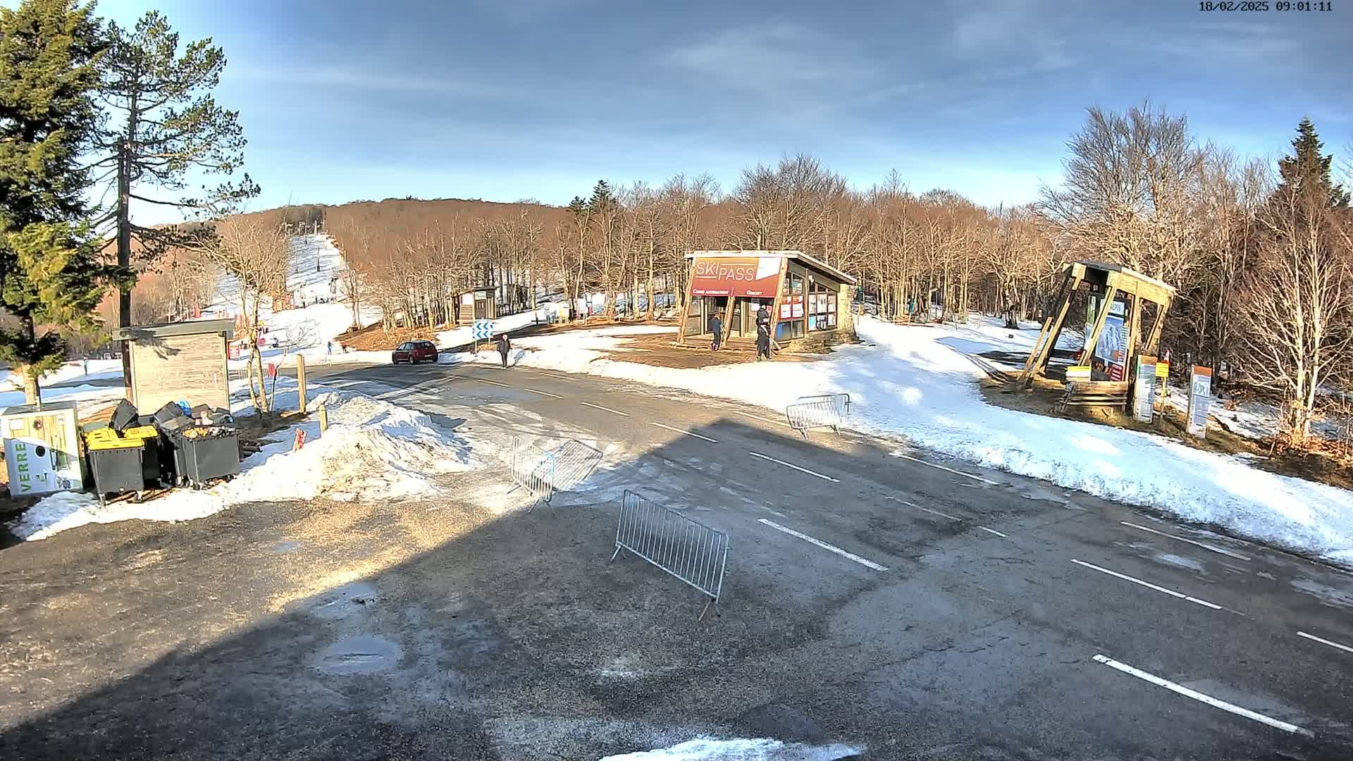 A sunny winter day at a ski resort shows a paved road with snow banks on either side, leading to a ski lift and a ski pass building; several people are visible near the buildings.