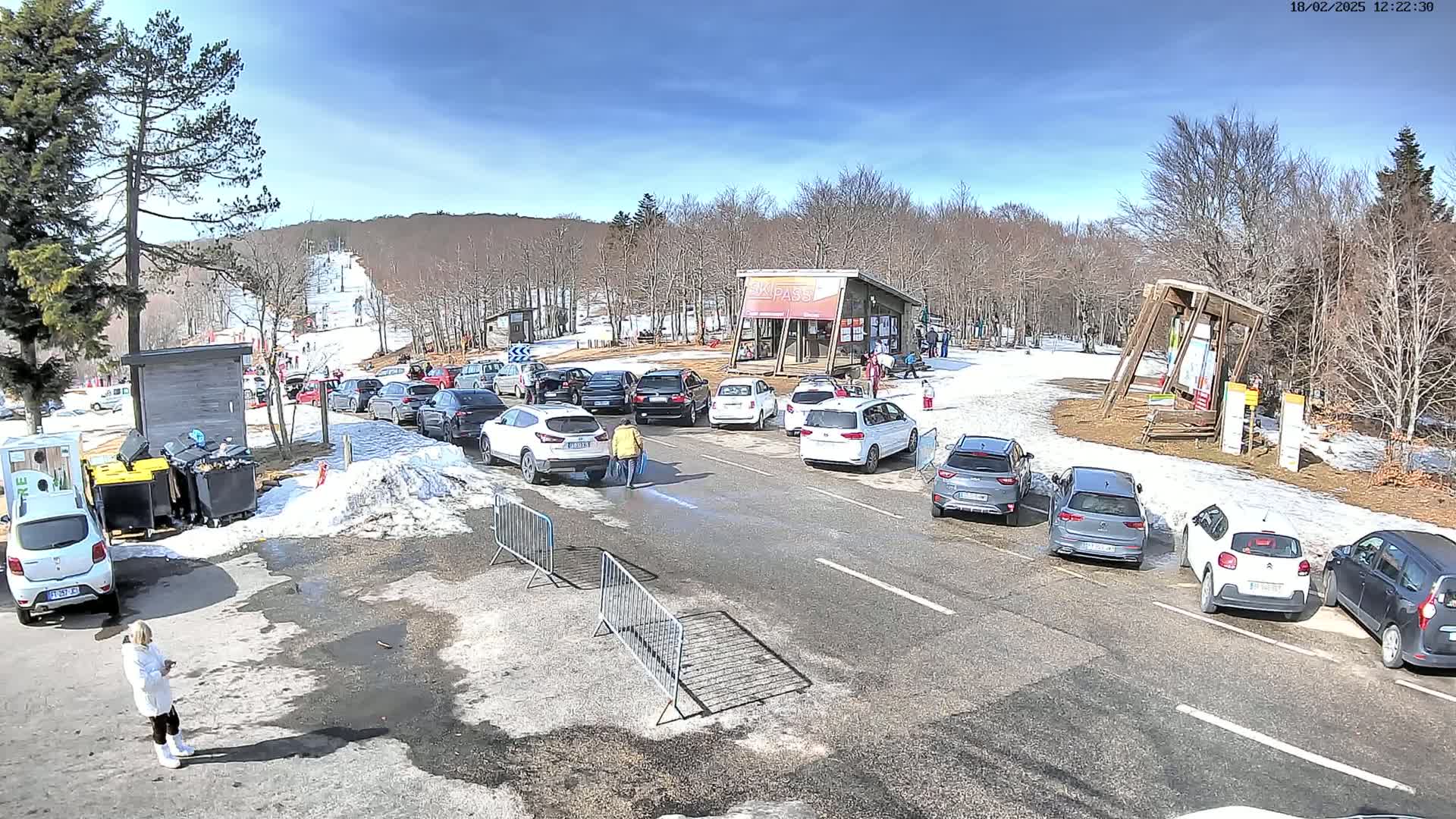 A parking lot with numerous cars is situated near a ski slope on a sunny day with patches of snow on the ground.