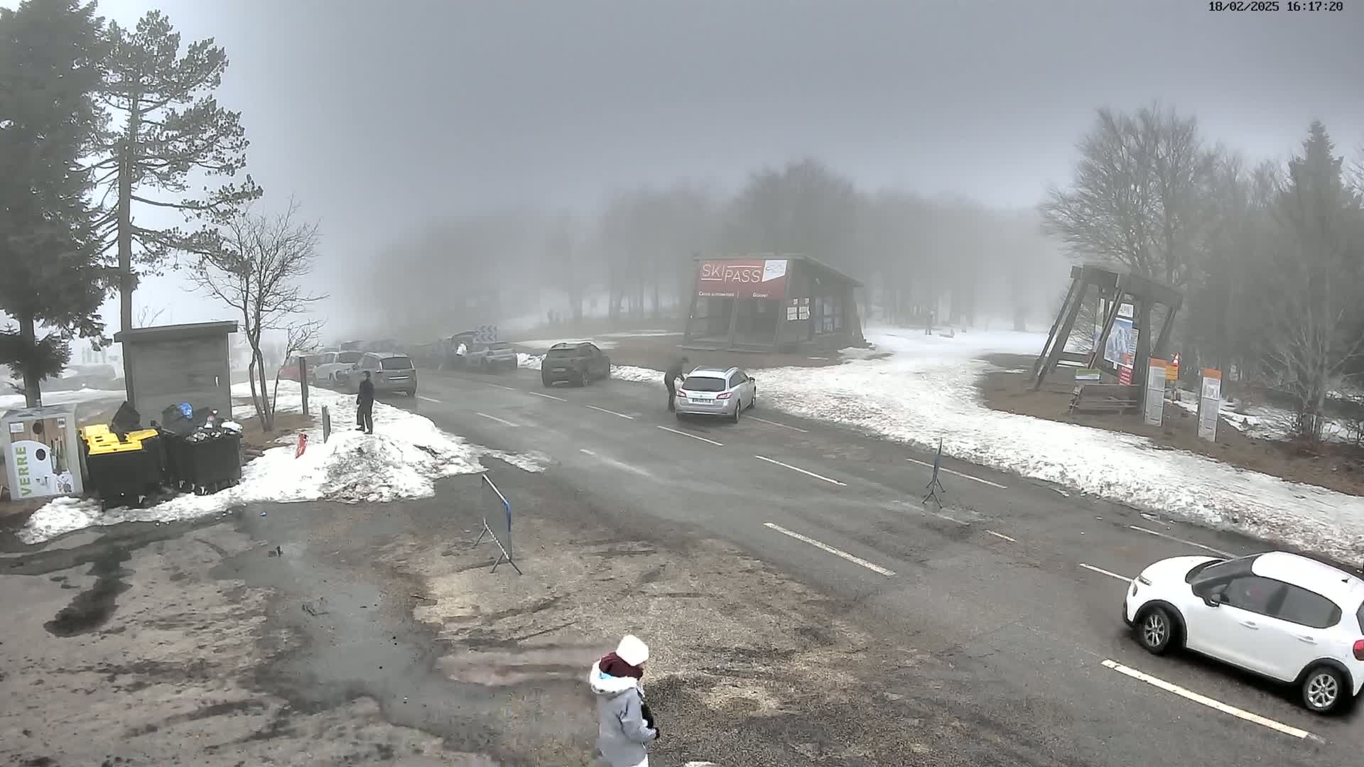 A foggy, snowy parking lot with several cars and a few people visible, near a ski pass building.