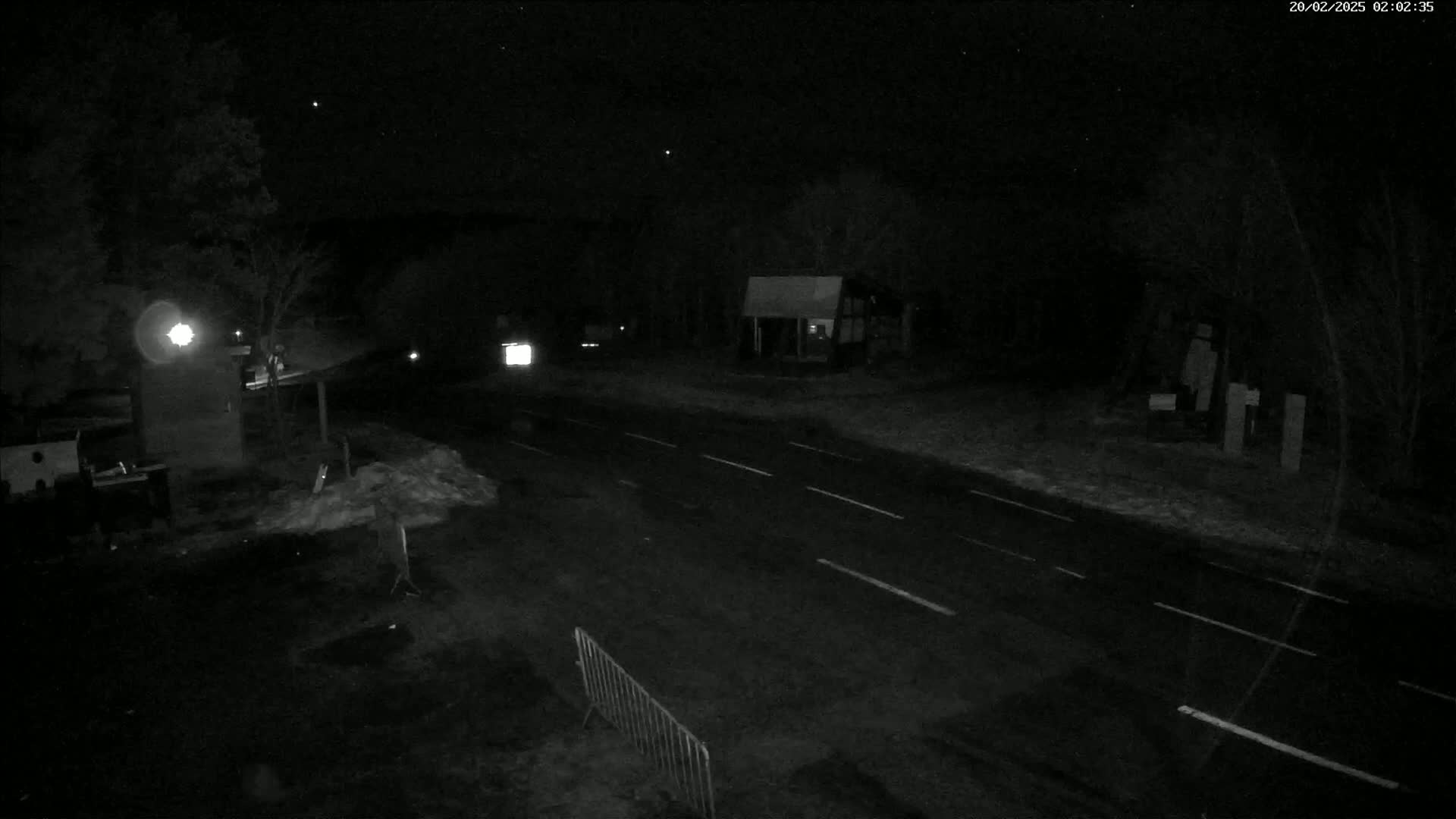 A nighttime, snow-dusted road scene shows a sparsely-lit roadway with a few vehicles and a small building under a dark, starry sky.