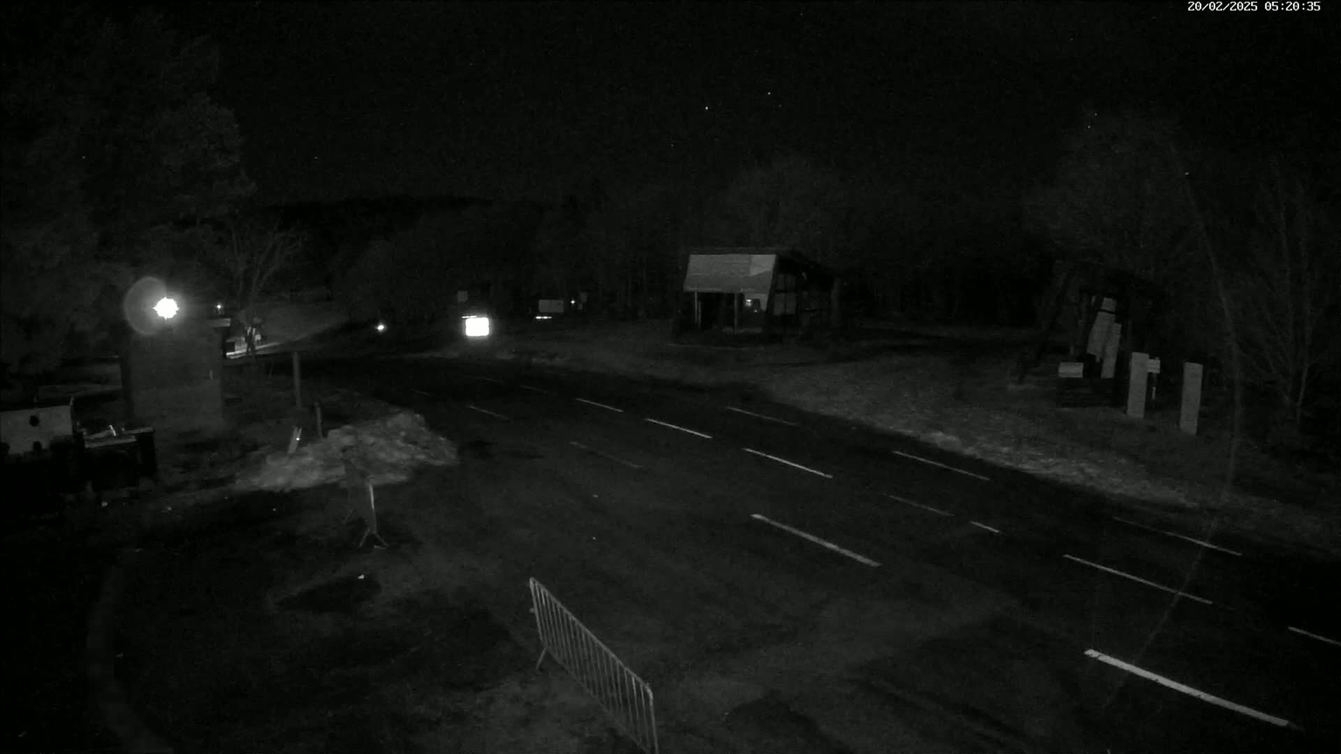 A dark, snow-dusted road at night is illuminated by a single streetlight, with a building and some trees visible in the background.
