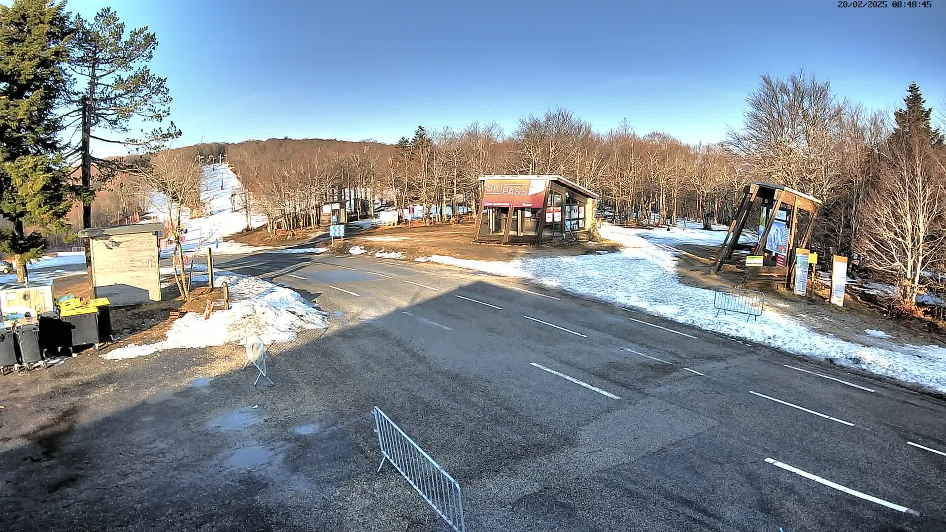 A paved road at a ski resort on a sunny day with patches of snow on the ground and in the background, surrounded by bare trees.