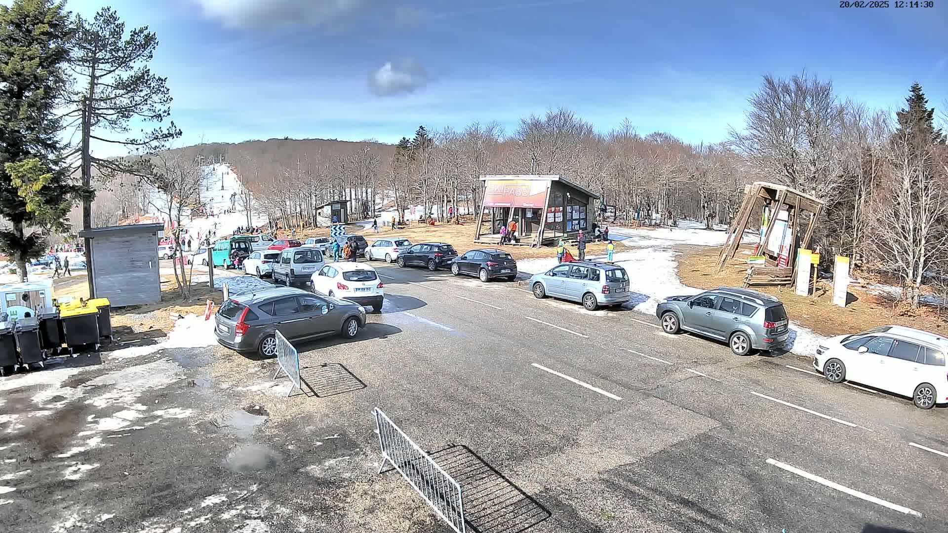 A sunny day at a ski resort shows a parking lot with numerous cars and people near a ski lift and a small building.
