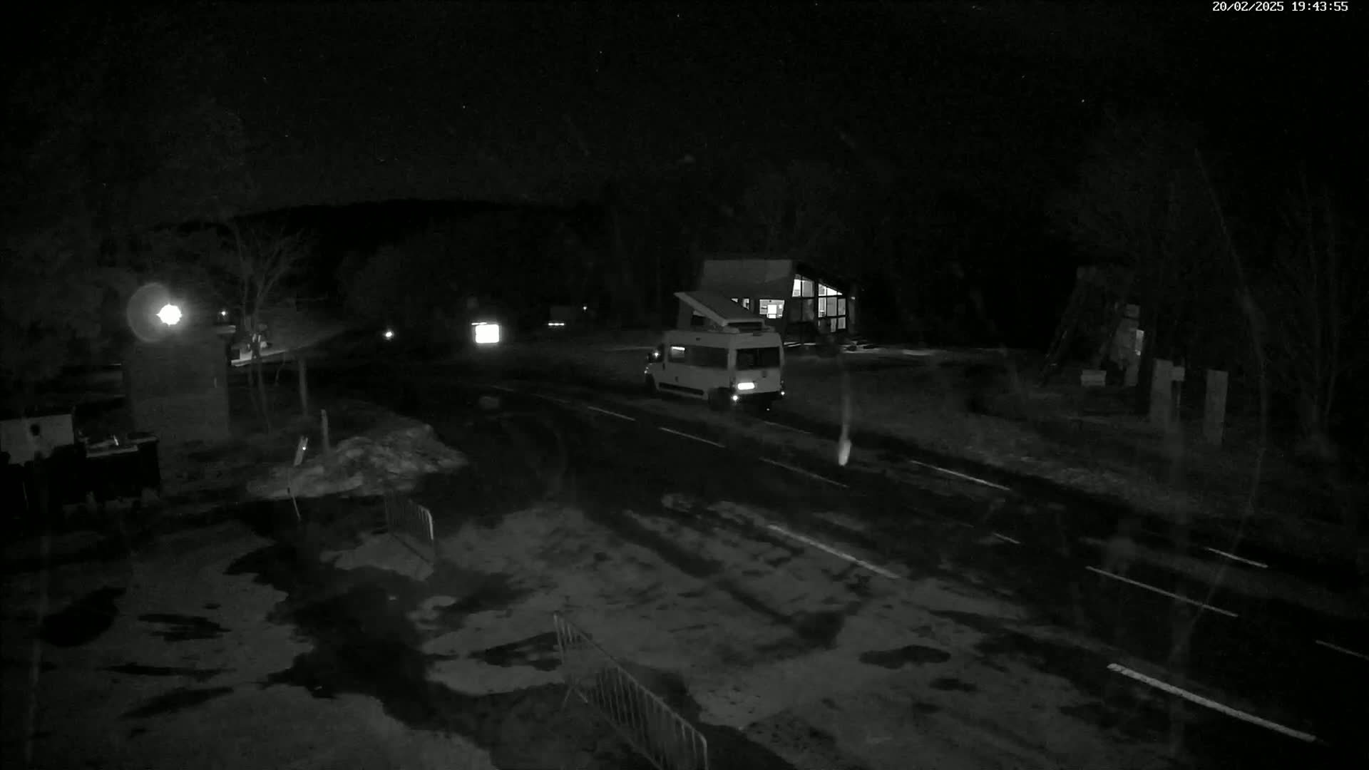 A nighttime, snow-dusted scene shows a camper van parked on a road near a building, with some other vehicles and objects visible in the low-light conditions.