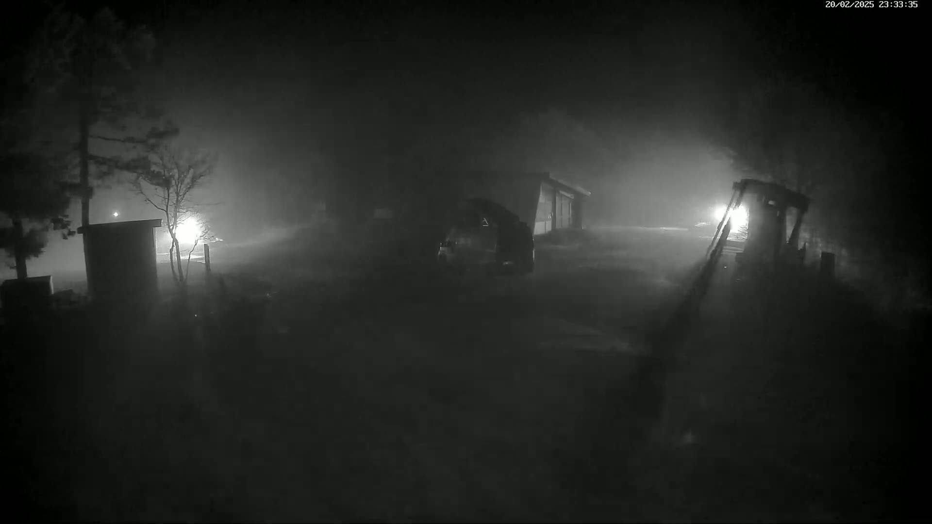 A nighttime, foggy scene shows a vehicle parked near a building and a piece of heavy machinery in a yard.