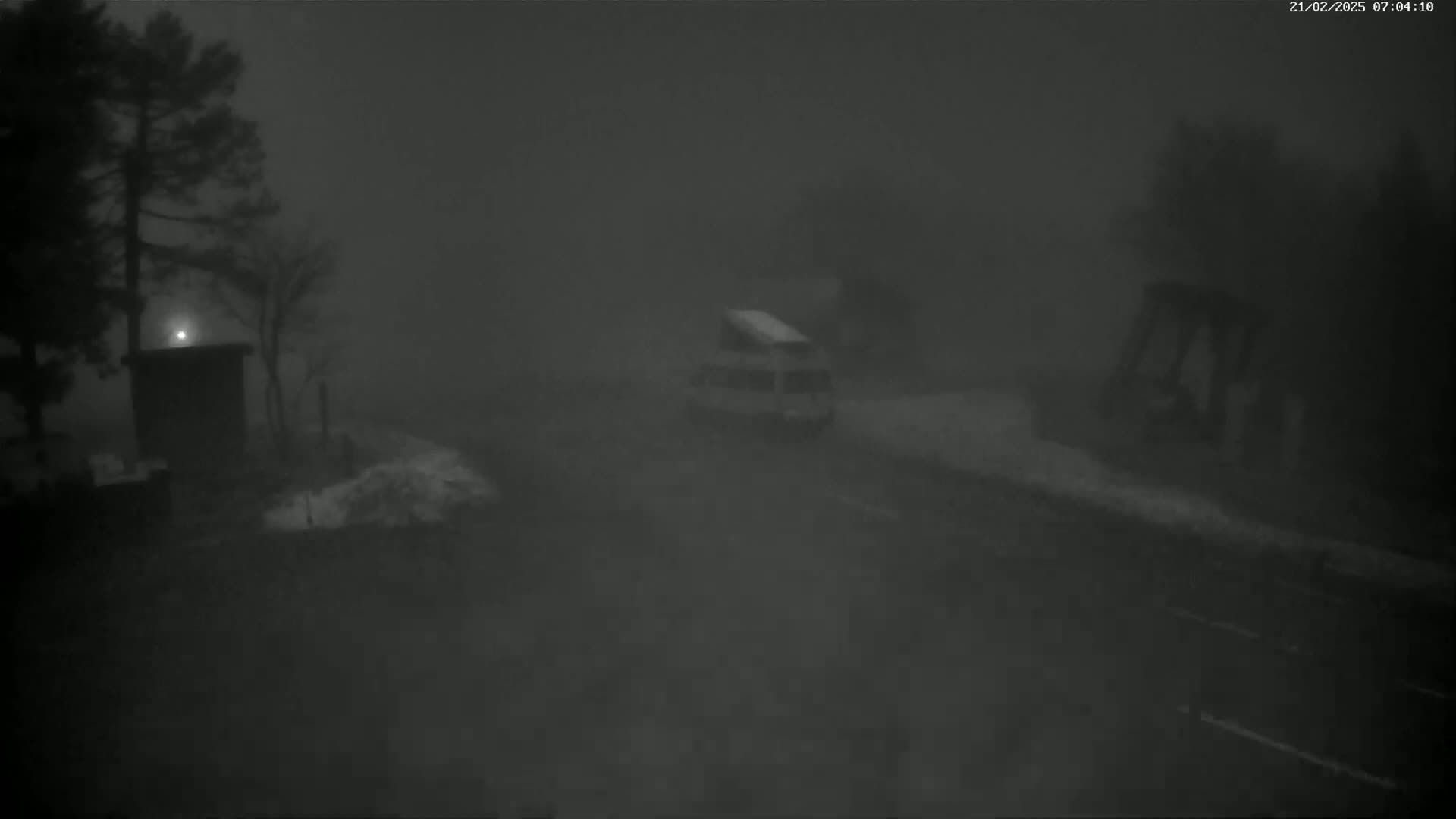 A vehicle drives down a road shrouded in heavy fog at night, past trees and a structure.