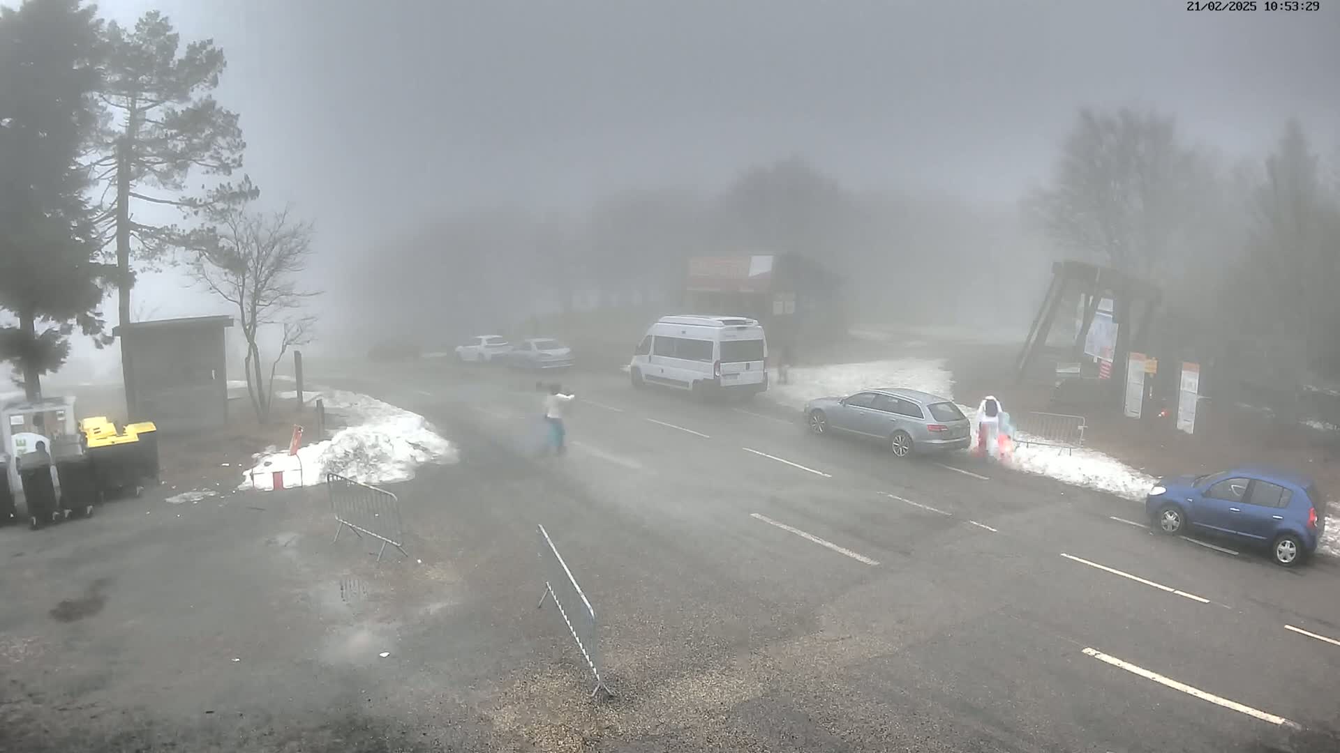 A foggy, snowy road scene shows several vehicles and pedestrians near a small building and snow banks.