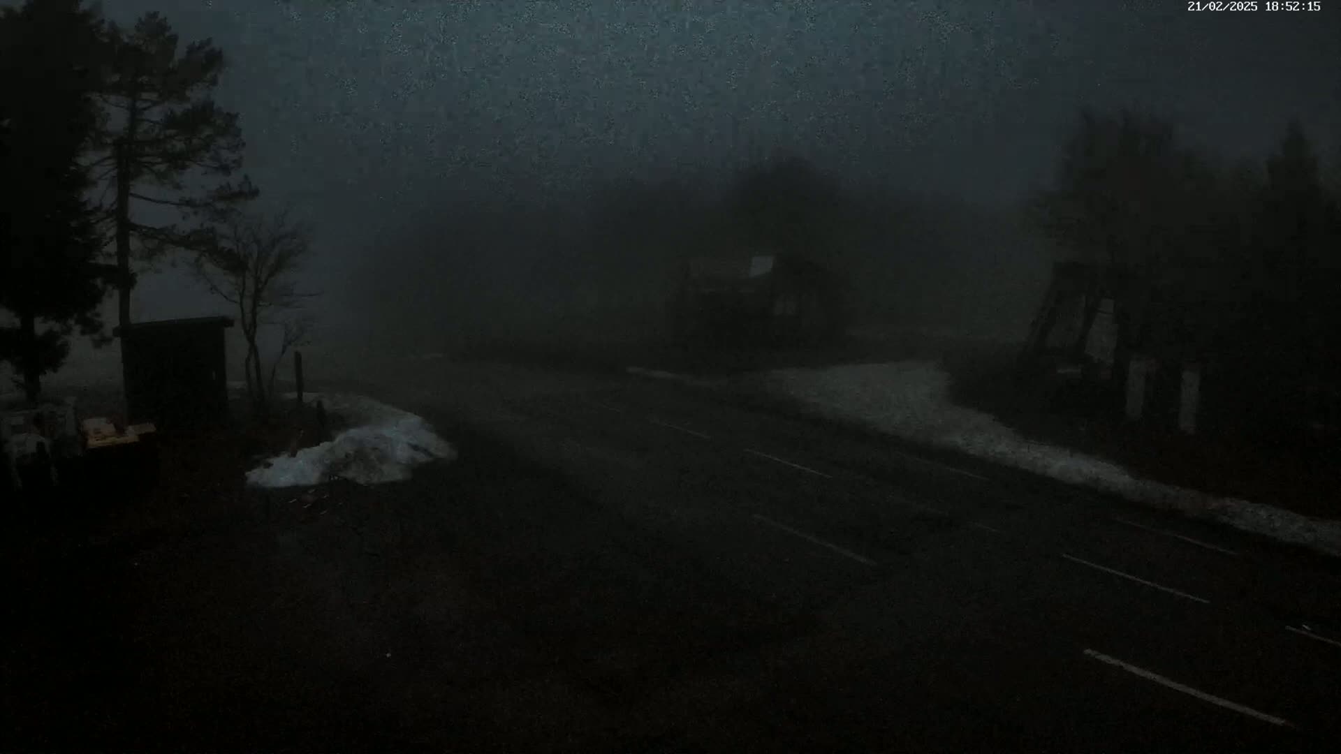 A dark, foggy road is seen with a few small structures and trees, some patches of snow are visible on the ground.