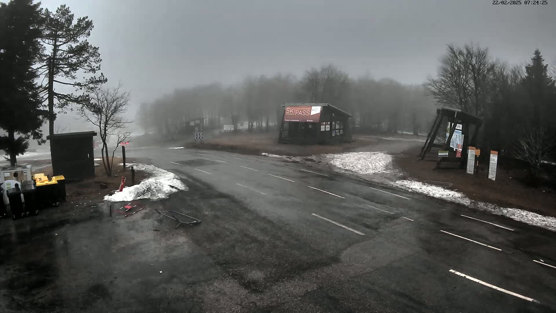 A foggy, wet road intersection near a ski pass building, with patches of snow and trees.