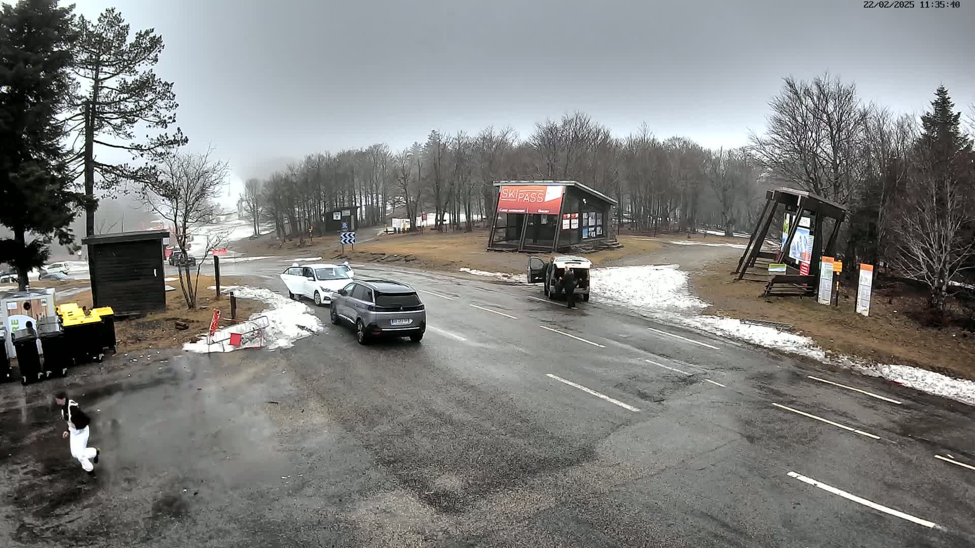 A wet, overcast day at a ski resort parking lot shows several cars, a small building, and some patchy snow on the ground.
