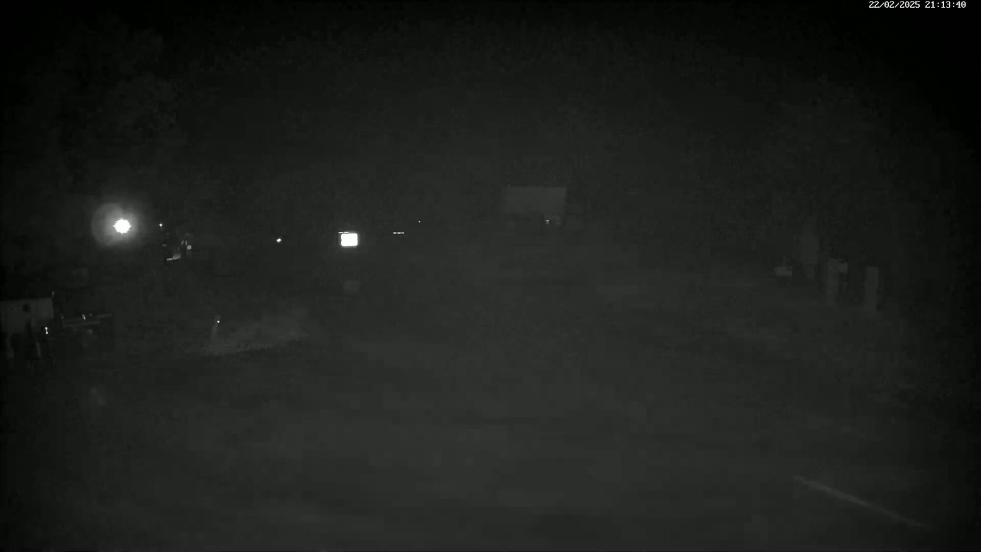 A dark, nighttime scene shows a dimly lit area with a few indistinct objects and vehicles.