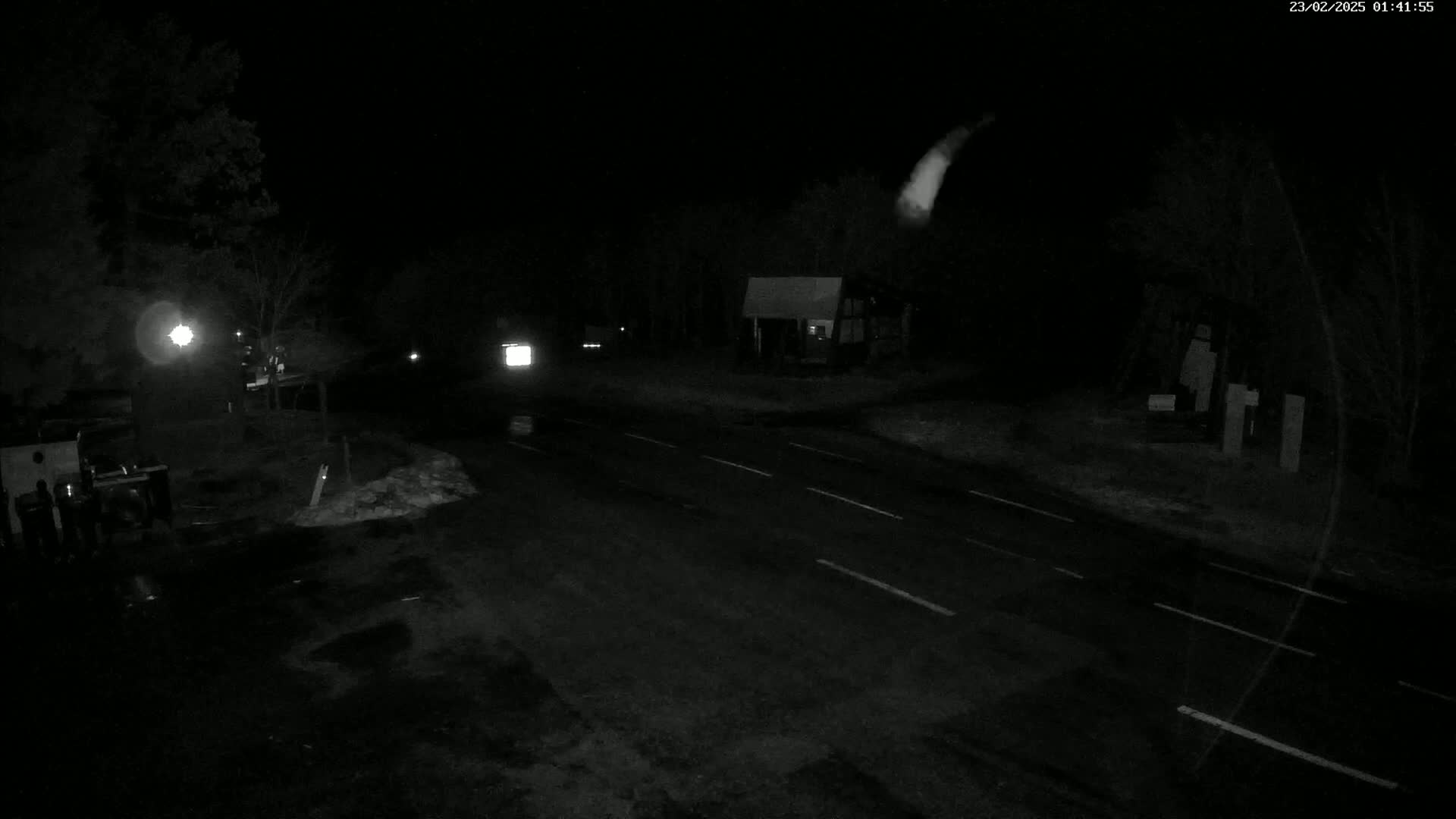 A dark, nighttime scene shows a road with a few vehicles, some buildings, and trees under a clear sky with a bright streak possibly a meteor.
