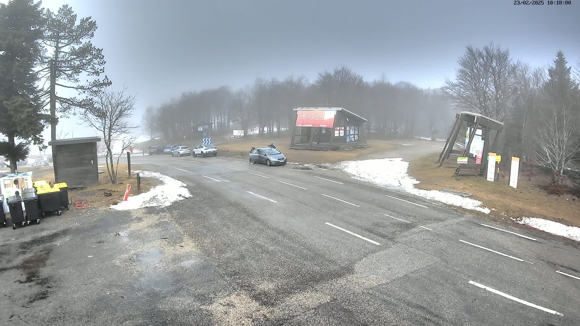 A foggy road scene shows several cars parked near a ski pass building and small patches of snow on the ground.