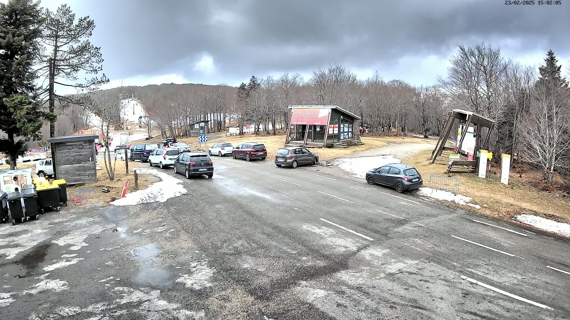 A parking lot with several cars is situated near a ski slope under a cloudy sky, with patches of snow on the ground.